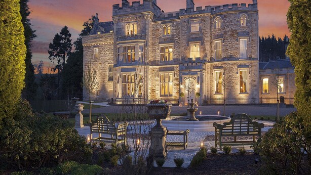 A large stone building with multiple windows and a central entrance, surrounded by a well-maintained garden with benches and a fountain. - Crossbasket Castle Hotel & Restaurant (Glasgow)