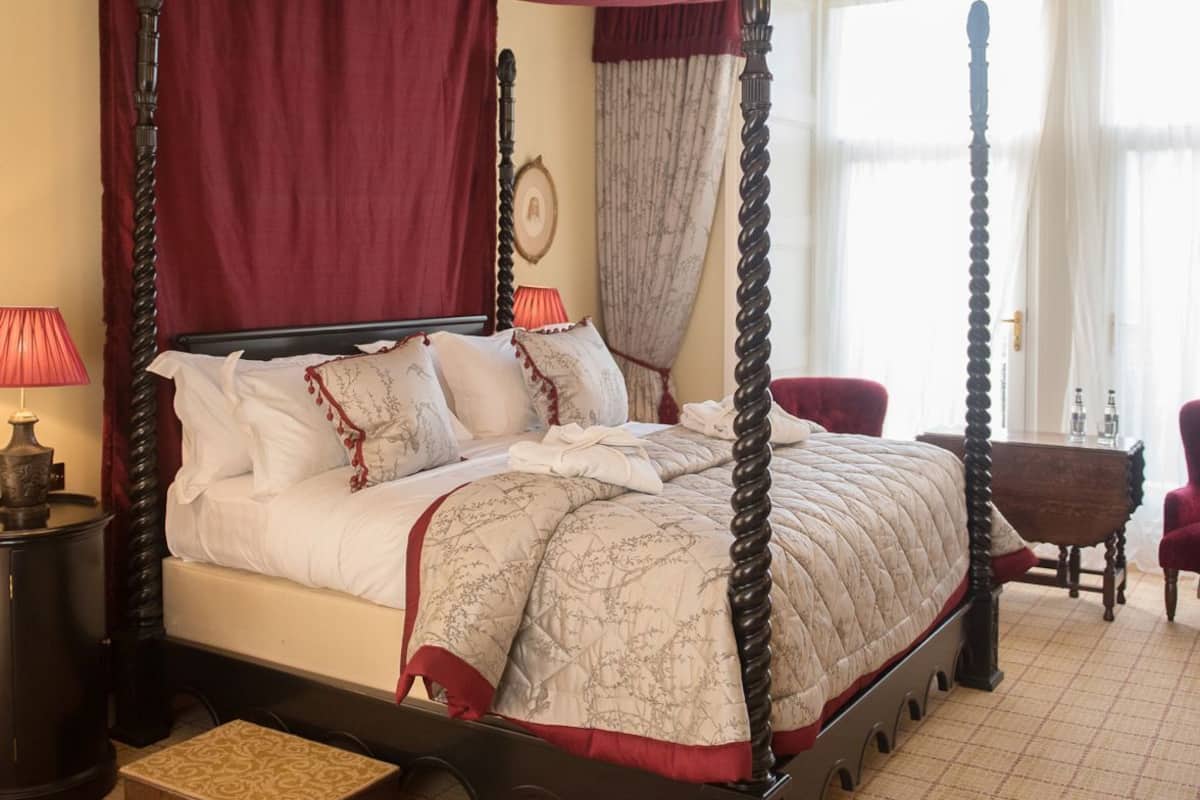 A four-poster canopy bed with a red canopy, a bedside table with a lamp, and a small wooden stool.