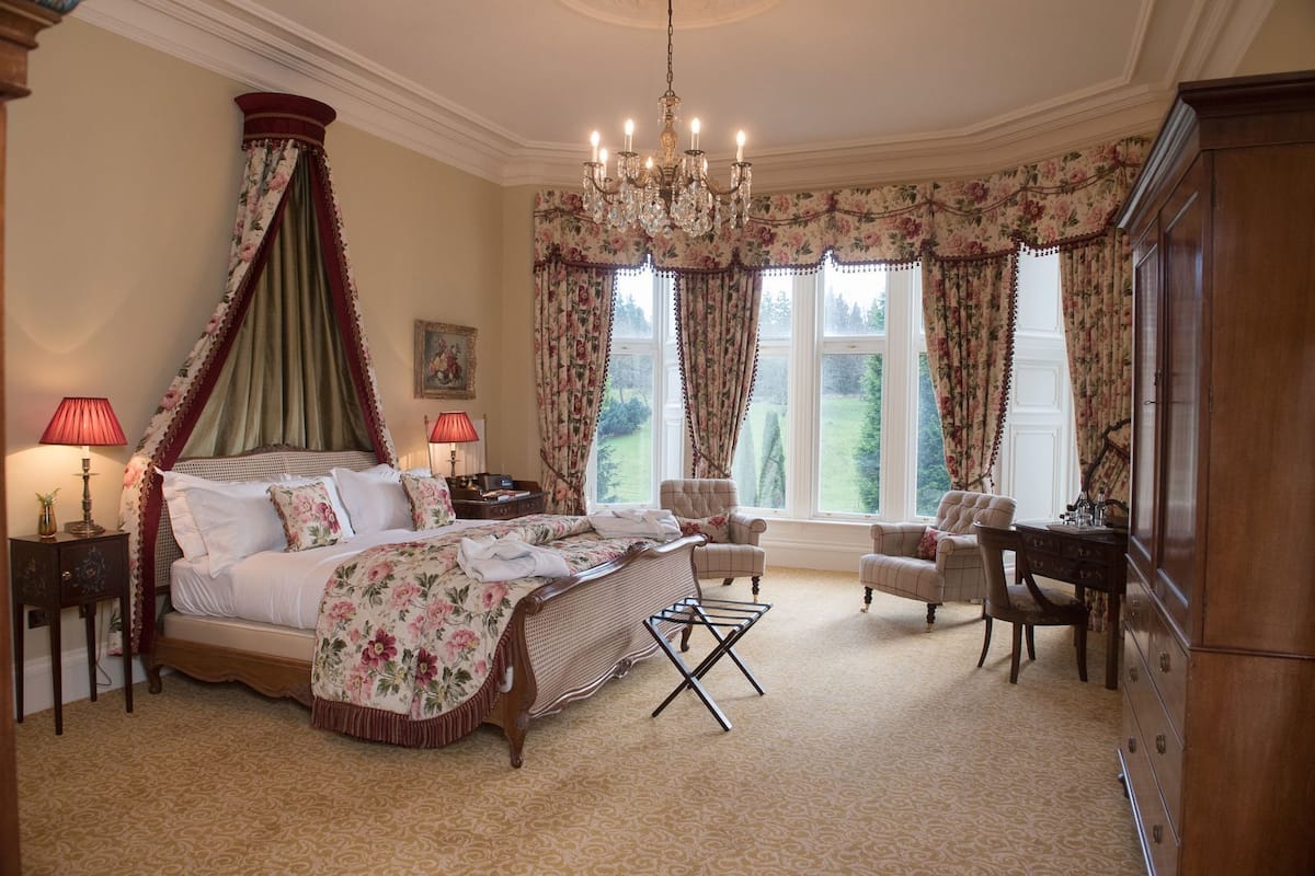 A bedroom with a four-poster bed, two armchairs, a desk, a chandelier, and large windows with floral curtains.