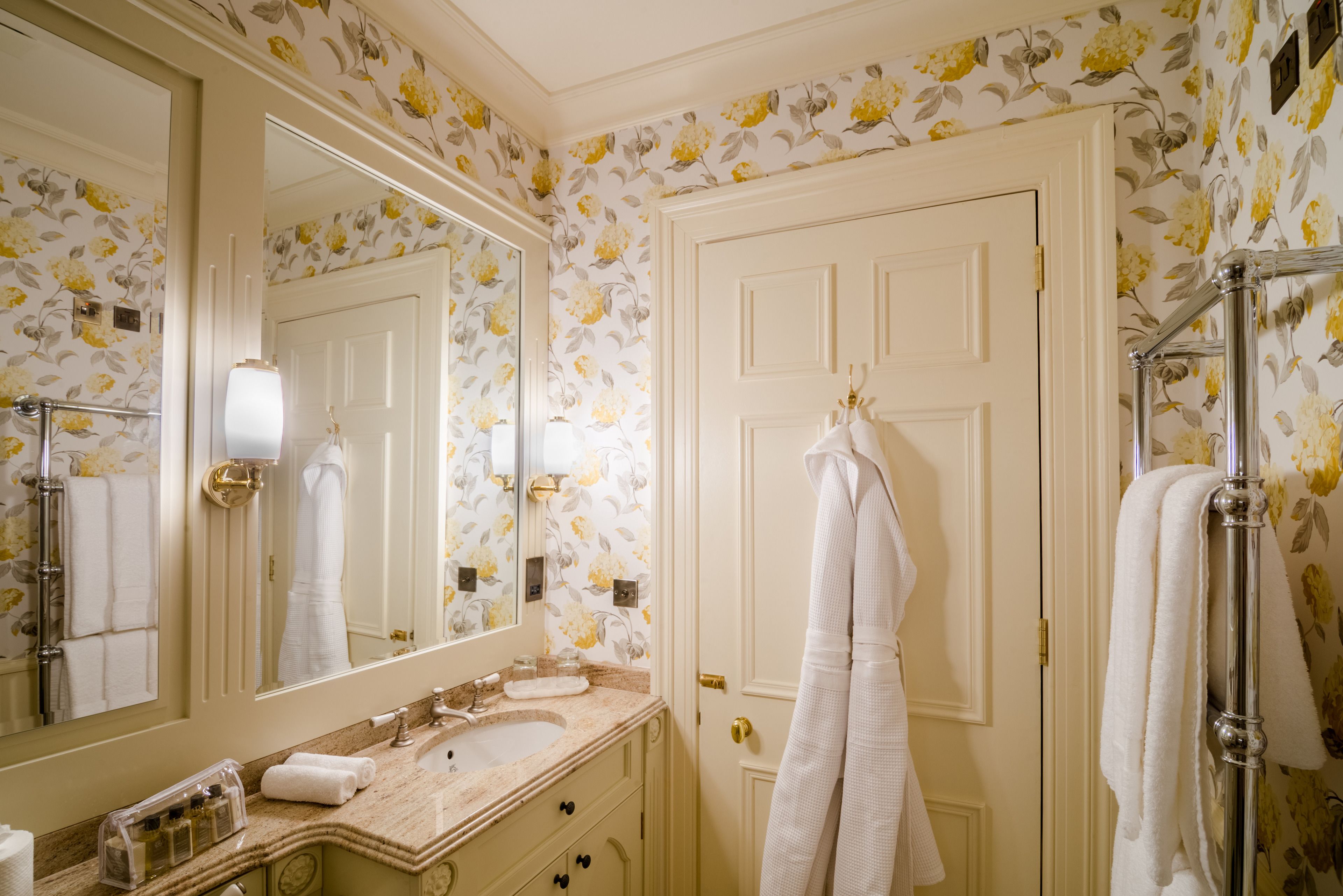 Un baño con lavamanos, espejo y puerta blancos, con papel tapiz floral y toallas blancas.