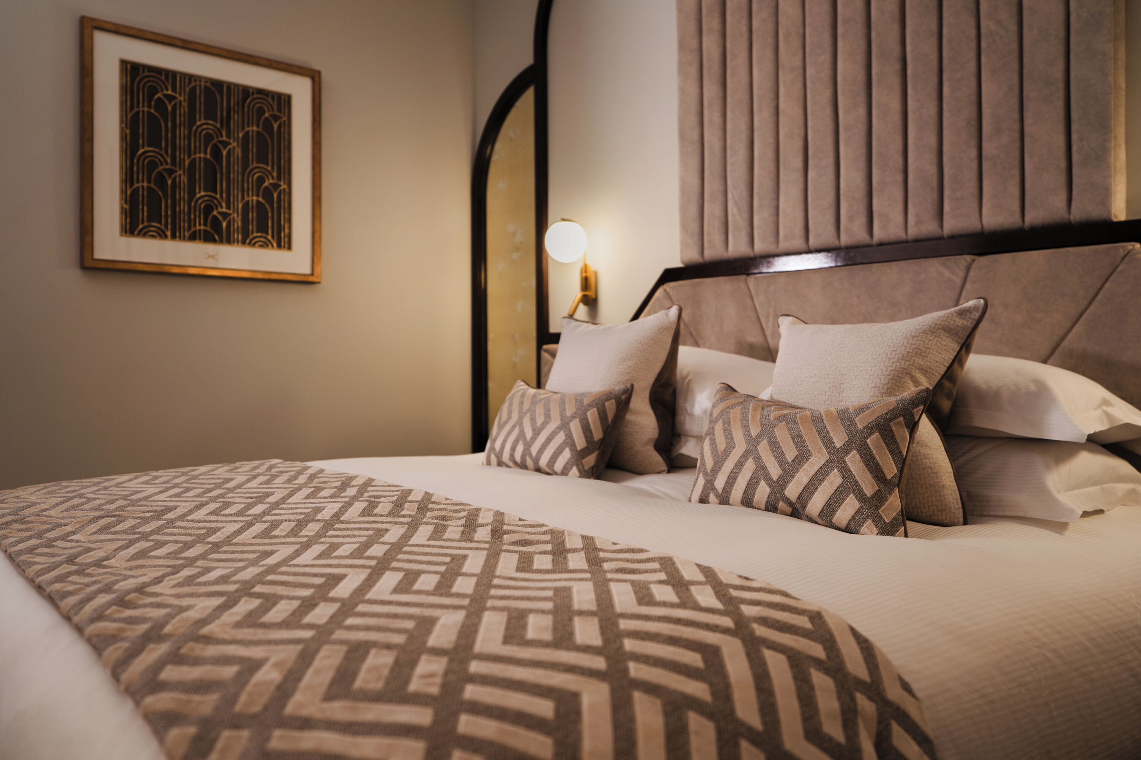 Una cama bien hecha con ropa de cama estampada, un cuadro colgado en la pared y una lámpara de noche.