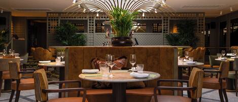 A restaurant with round tables, brown upholstered booths, and potted plants.