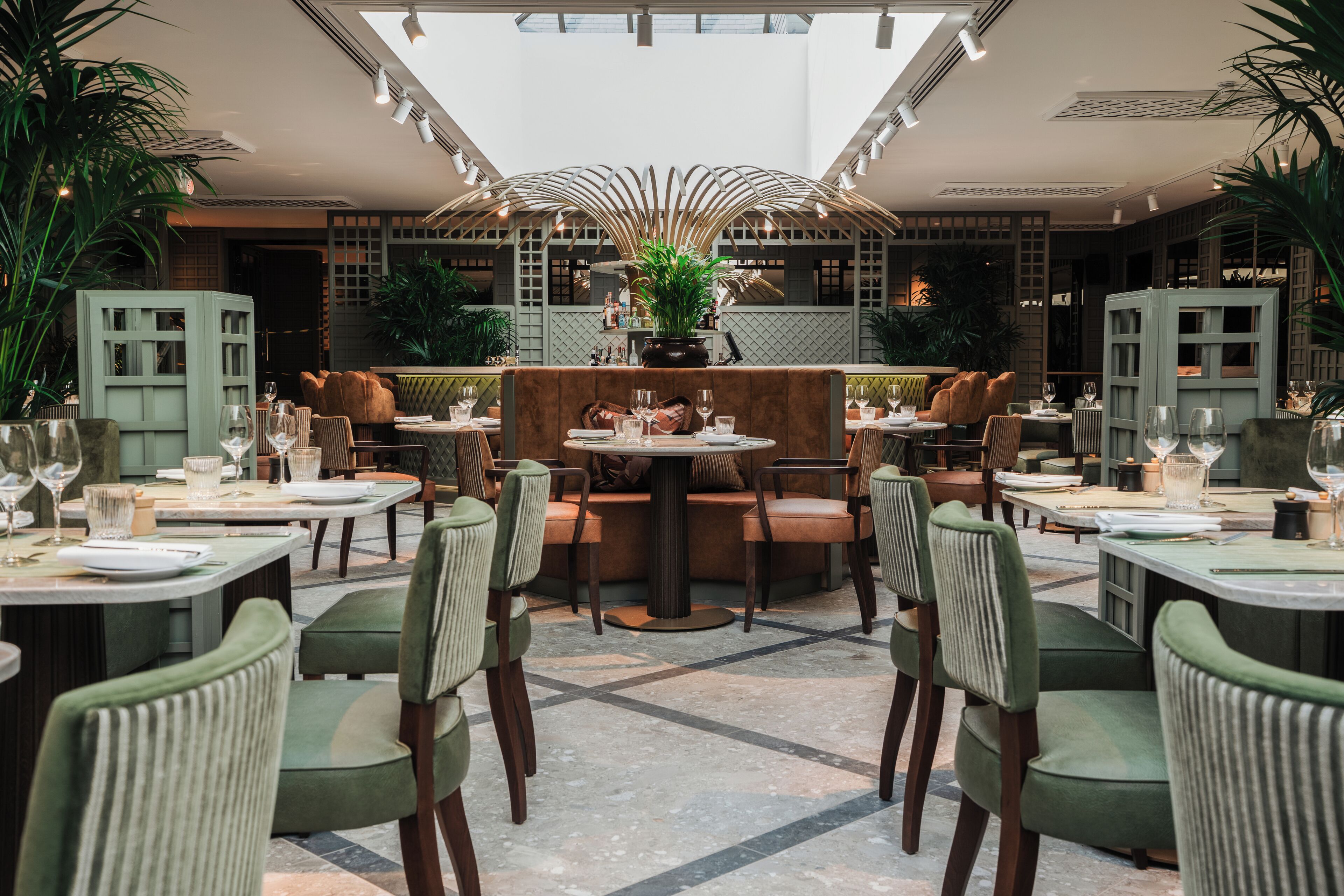 Um restaurante com várias mesas e cadeiras, uma área de estar central com uma mesa redonda e plantas em vasos.