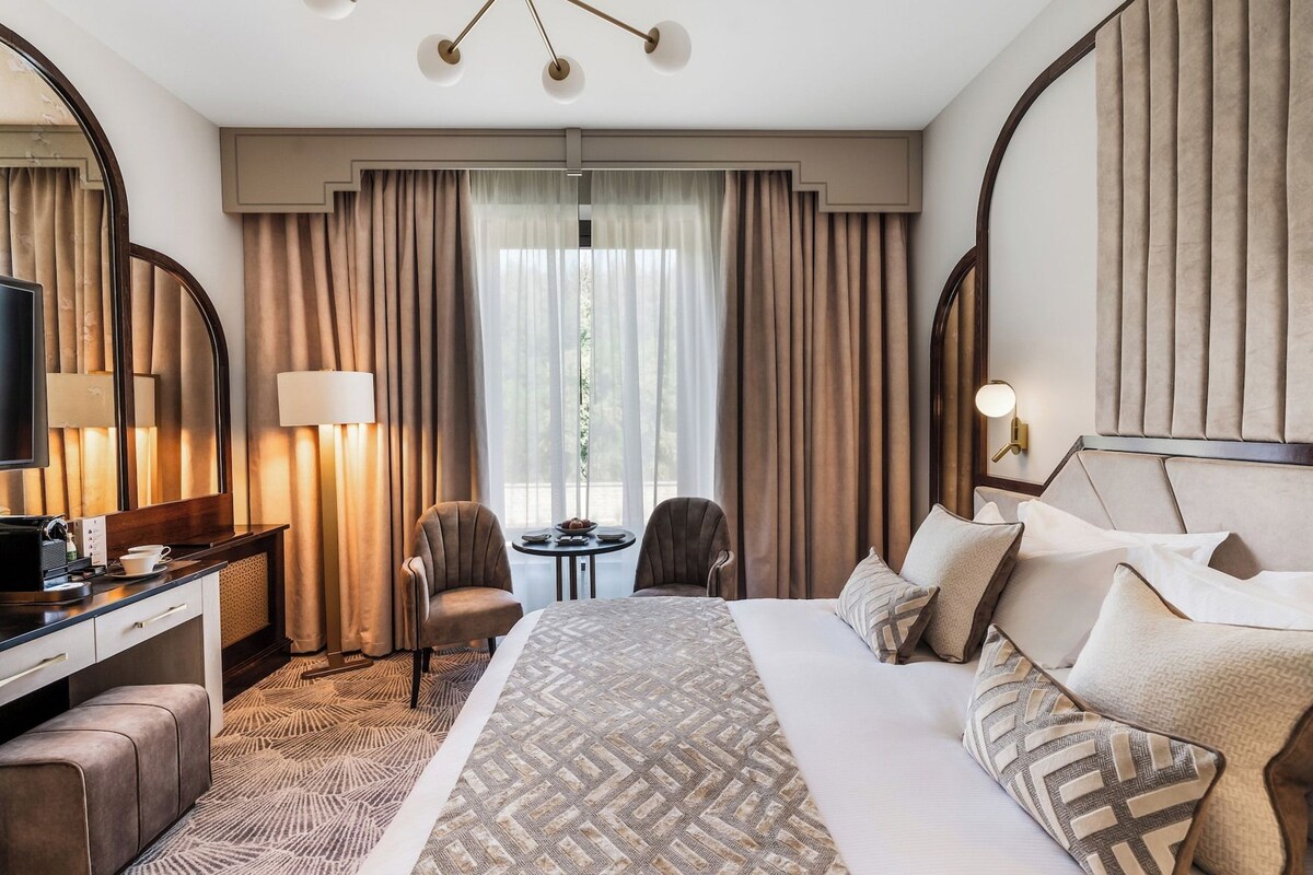 Habitación de hotel con una cama grande, dos sillones, un escritorio con televisor y vistas al exterior por la ventana.