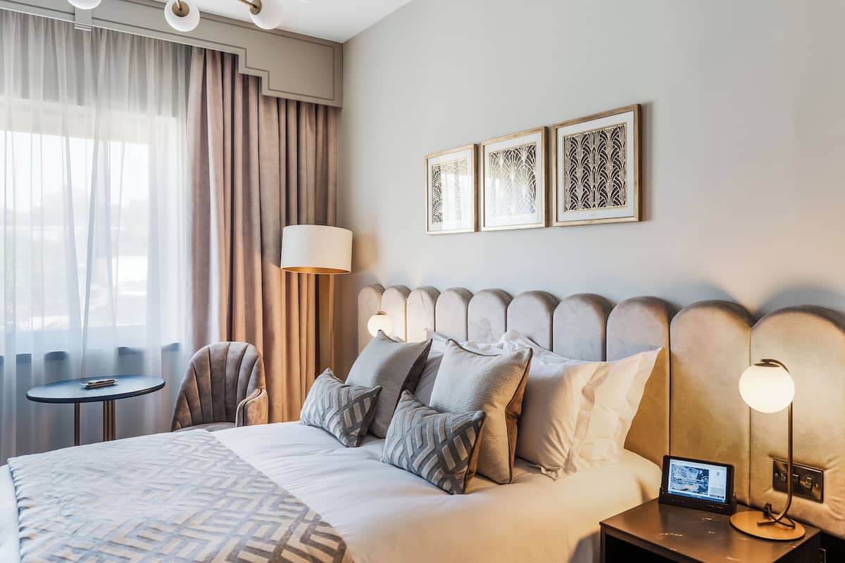 A hotel room with a large bed, a bedside table with a lamp, a chair, and a small table. There are curtains, a wall-mounted light, and three framed pictures on the wall.