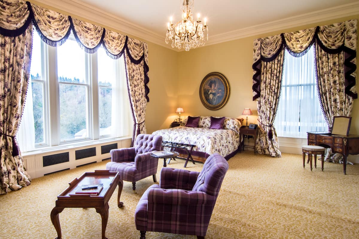 A room with a large bed, two armchairs, a small table, a dresser, and a chandelier.