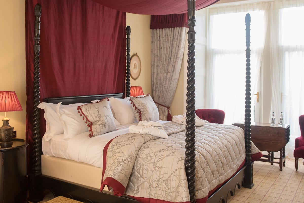 A four-poster canopy bed with a red canopy, a wooden headboard, and a footboard. There are two bedside tables with lamps, a small wooden stool, and a chair. The room has a checkered floor and a window with curtains.