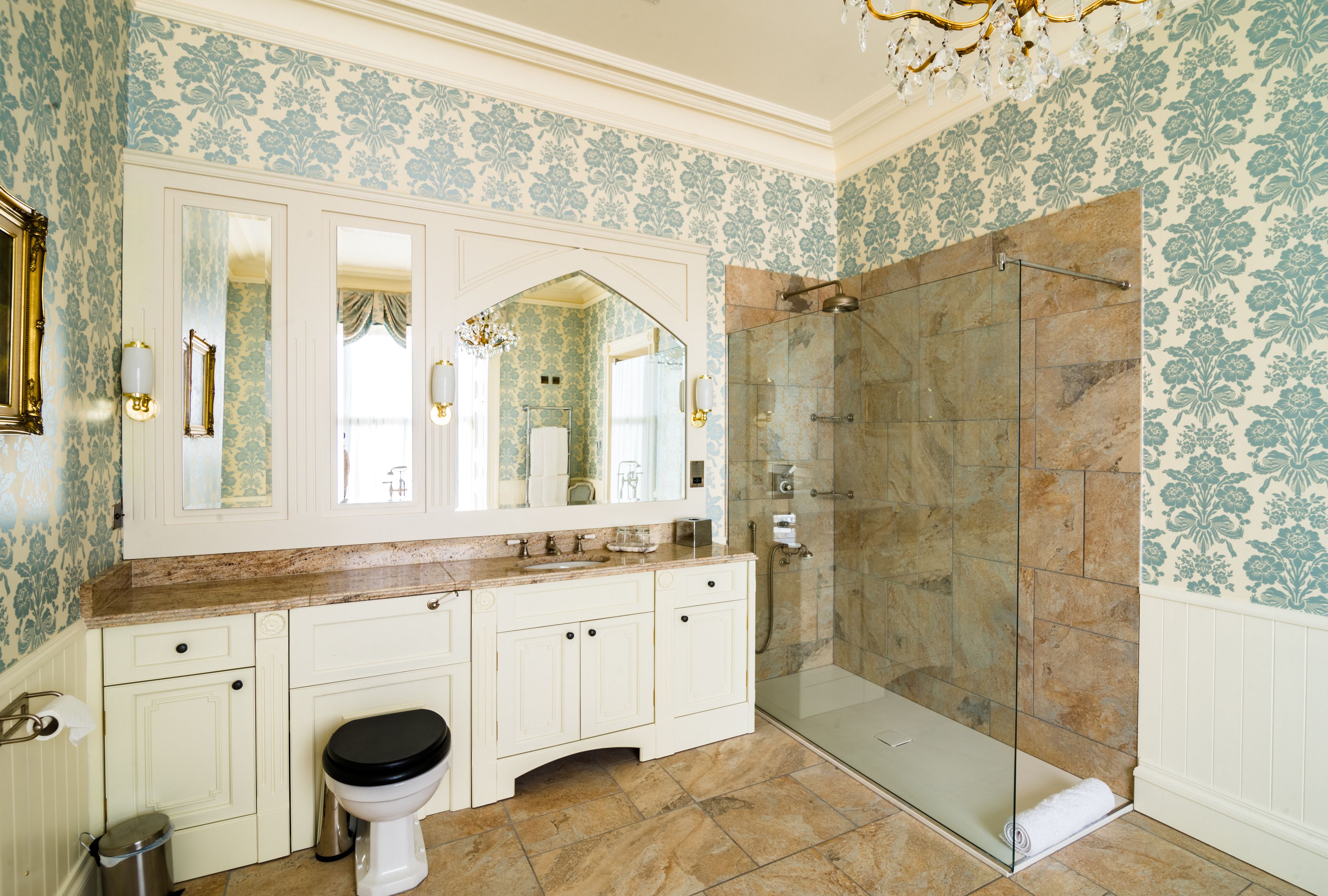 A bathroom with a large mirror, white cabinetry, a toilet, a shower with glass doors, and patterned wallpaper.