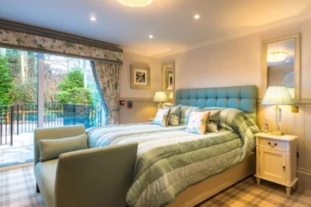 A bedroom with a large bed, a nightstand, a lamp, and a view of the outdoors.