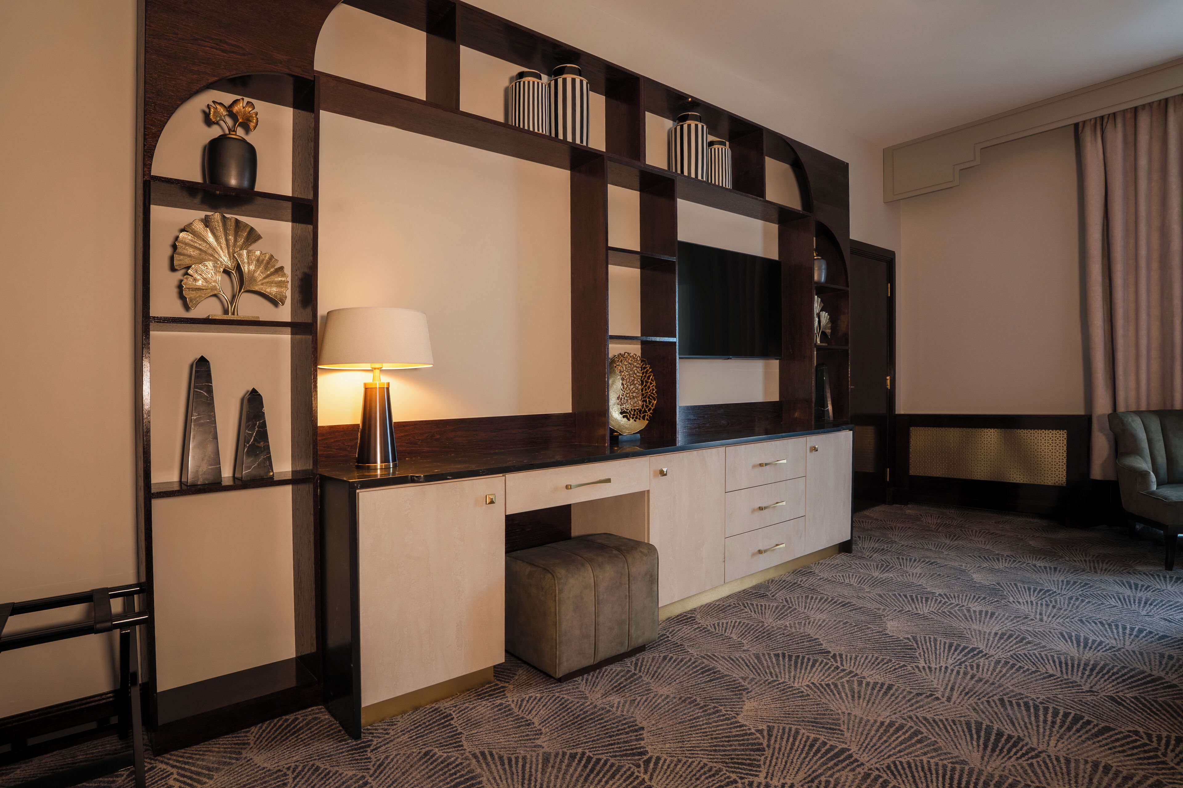 A modern hotel room with a large desk, a mirror, a lamp, and a patterned carpet.