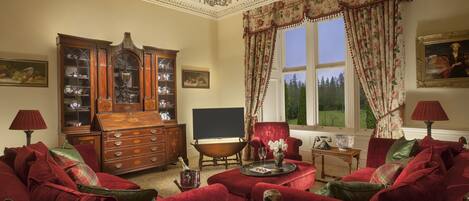 A traditional room with a large wooden cabinet, a red armchair, a sofa, a fireplace, and a view of a forest through a window.