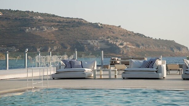 Un espace aménagé au bord de la piscine, avec des chaises longues et des tables, offrant une vue sur un plan d’eau et une colline parsemée de bâtiments.