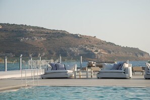 Un espace aménagé au bord de la piscine, avec des chaises longues et des tables, offrant une vue sur un plan d’eau et une colline parsemée de bâtiments.