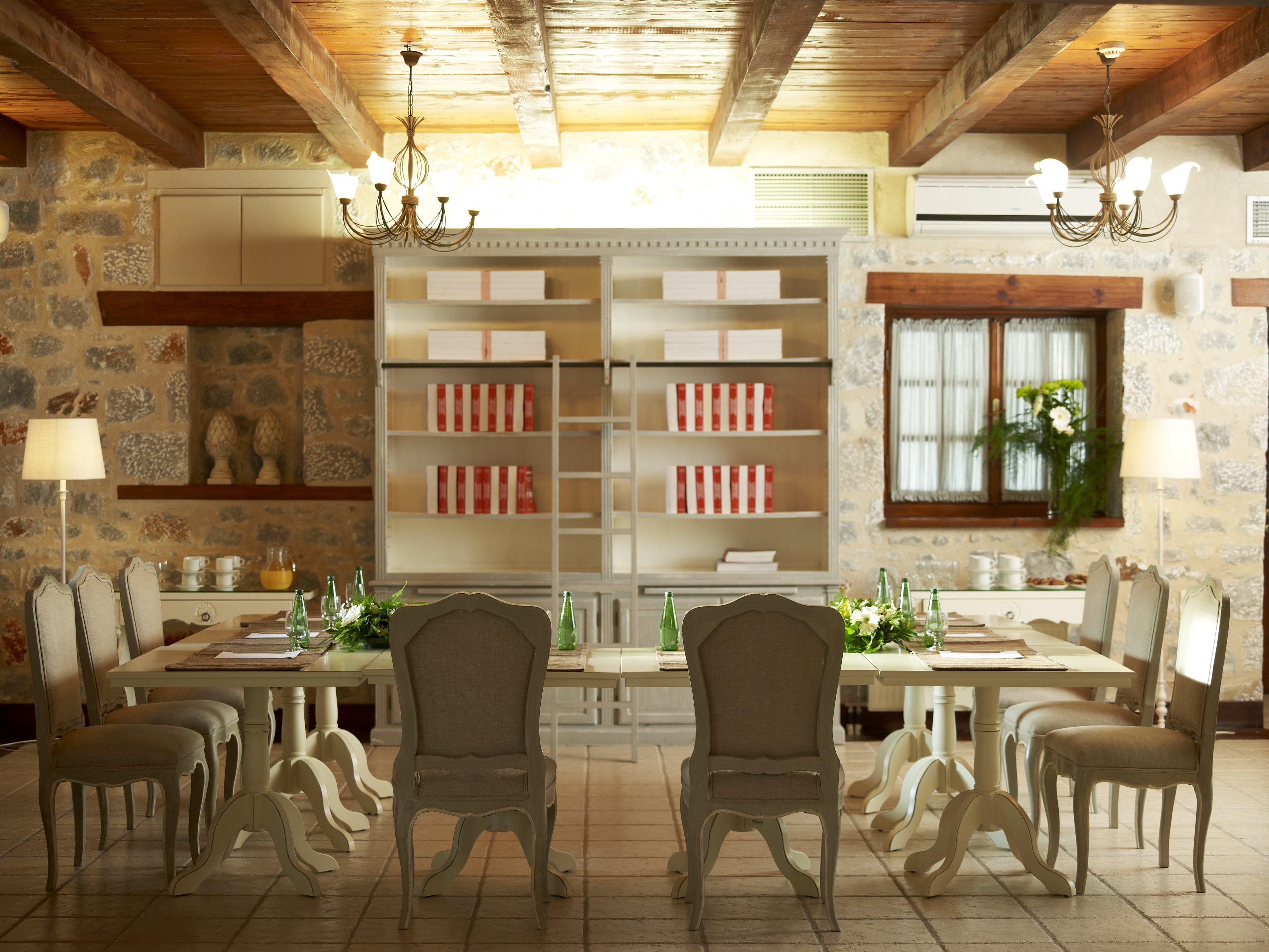A dining area with a large table, multiple chairs, and a chandelier. There are shelves with books and decorative items, and a window with a plant.