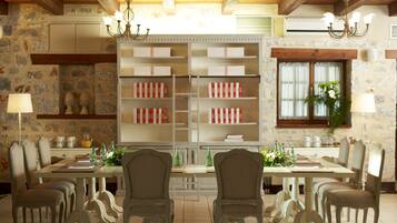 A dining area with a large table, multiple chairs, and a chandelier. There are shelves with books and decorative items, and a window with a plant.