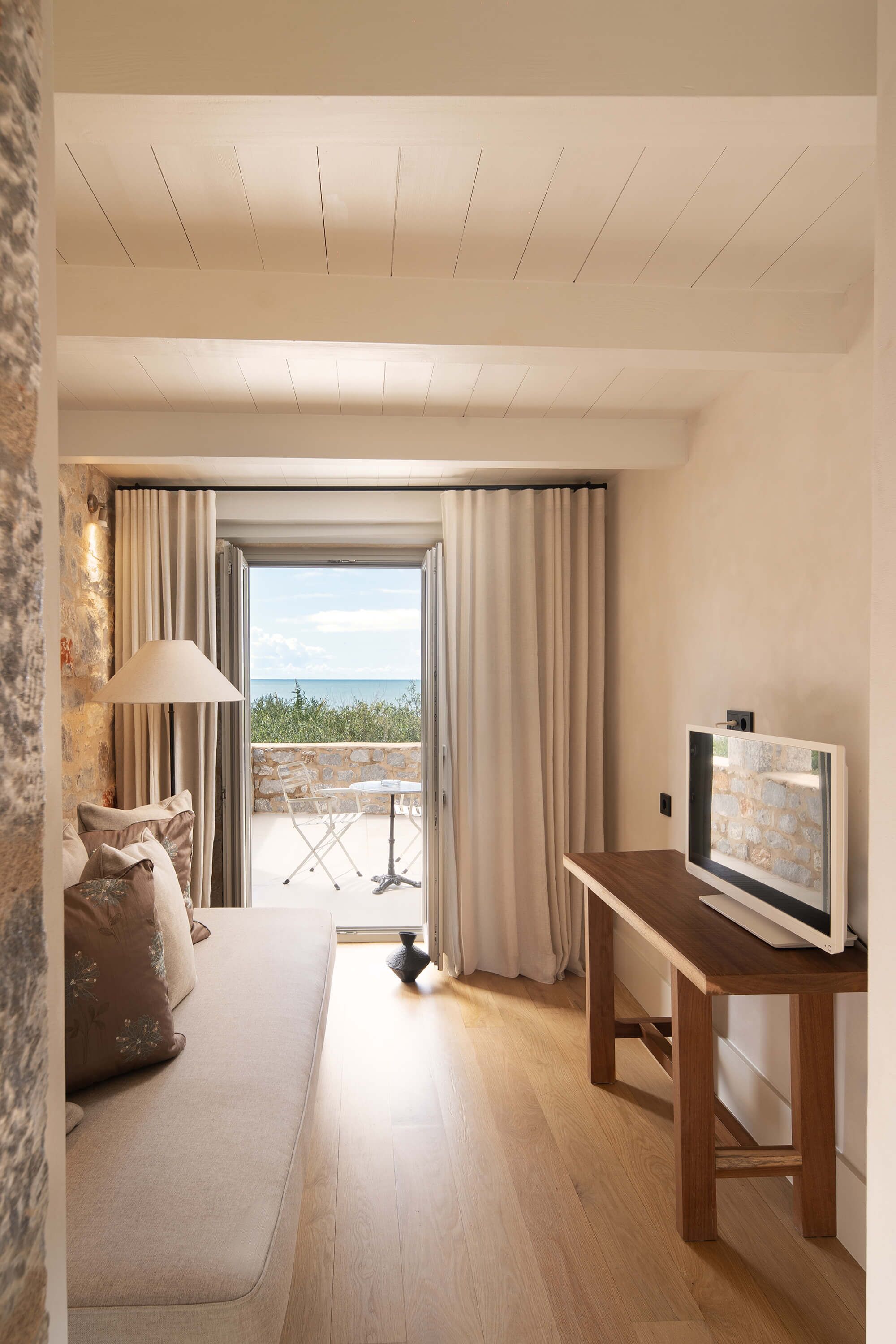 A room with a sofa, a TV on a wooden stand, a stone wall, and a view through a glass door.