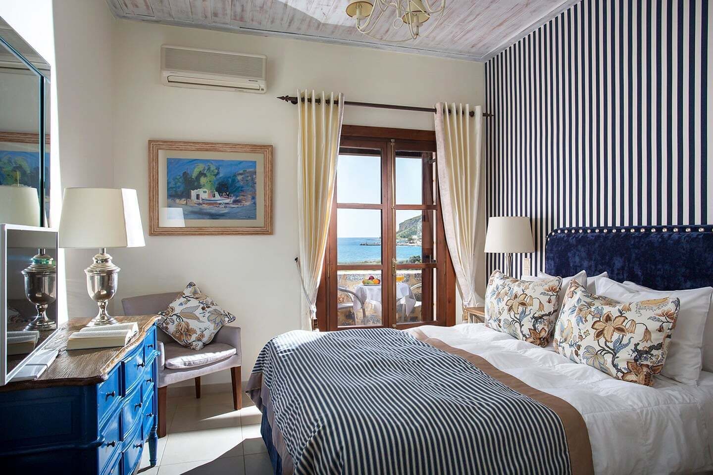 Une chambre d’hôtel avec un lit, un bureau doté d’une lampe, une chaise, un tableau, une fenêtre avec des rideaux et une vue sur la mer.