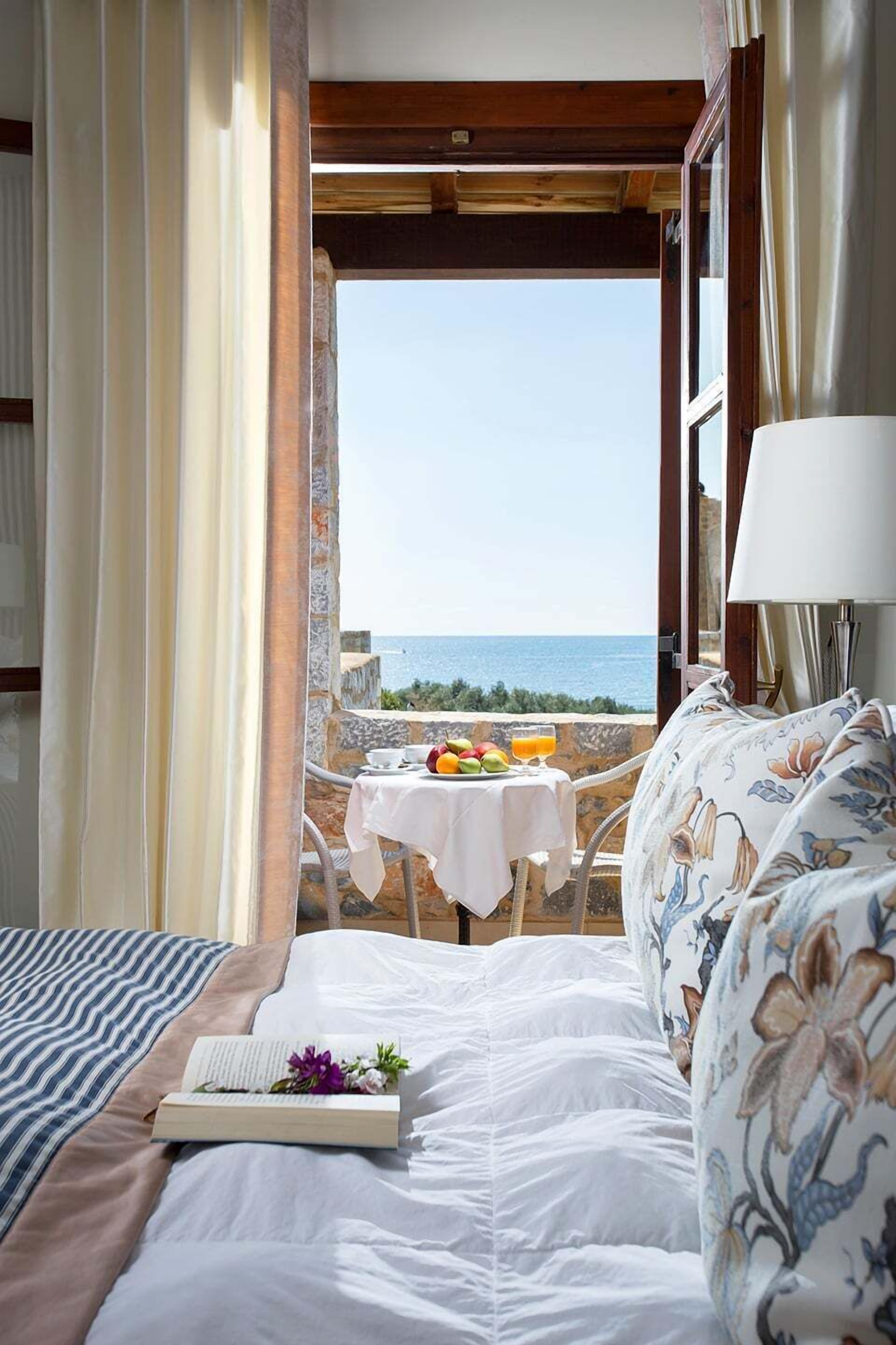 A bedroom with a bed, a bedside table with a book and flowers, a view of the sea, and a table with fruits and a glass of orange juice.