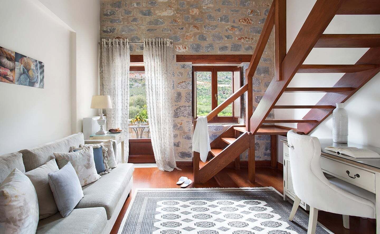 A cozy room with a wooden staircase, a sofa, a desk, and a view of the outdoors.