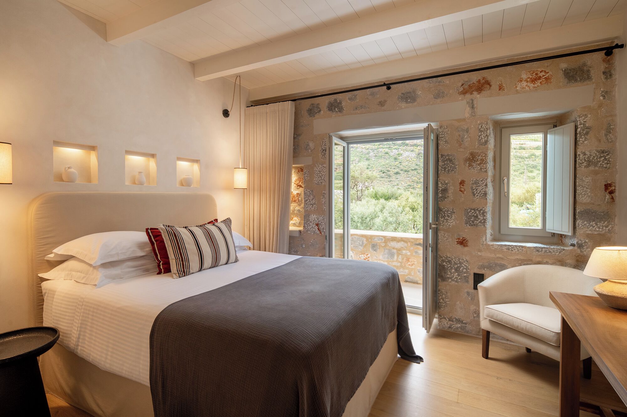A bedroom with a bed, a chair, a table, and a view of the outdoors.