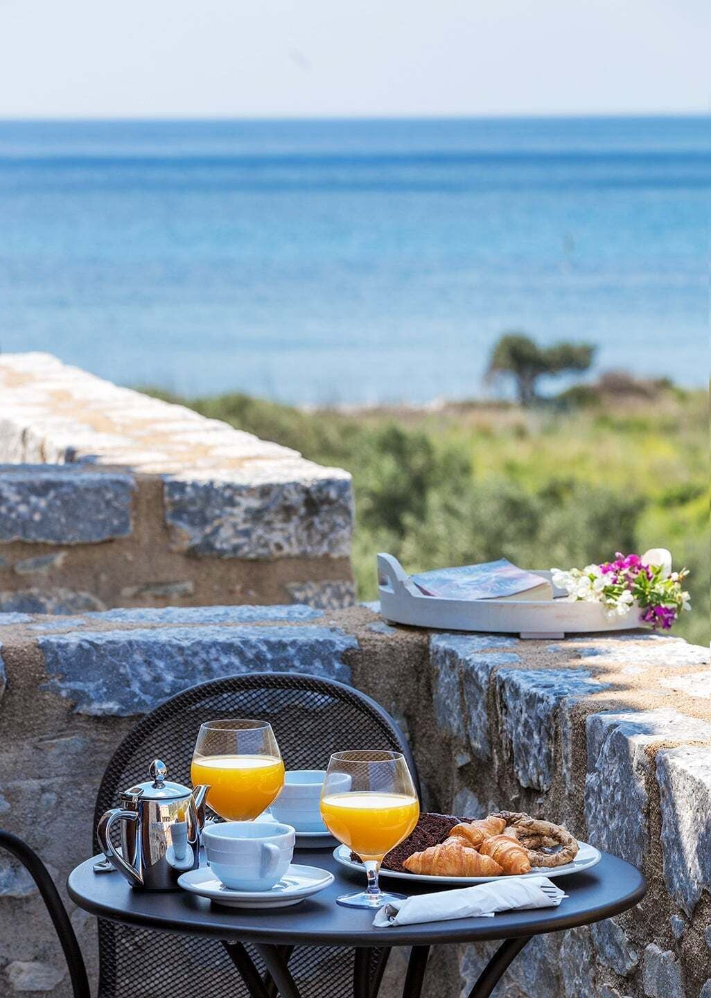 A table with breakfast items, a view of the sea, and a stone wall.