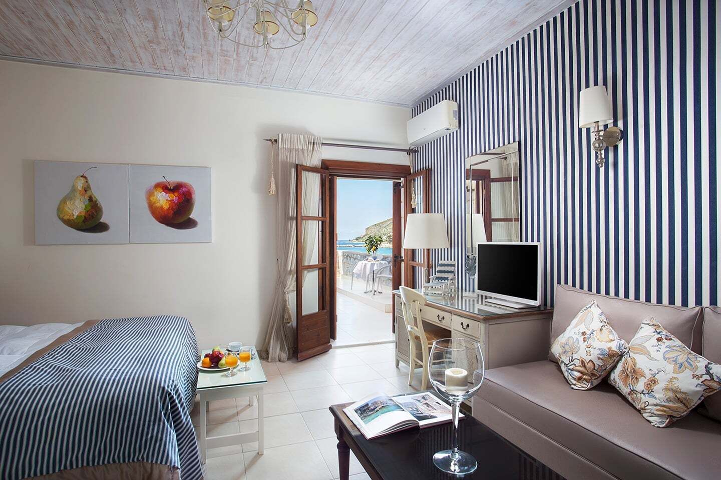 A hotel room with a bed, a desk with a computer, a sofa, a dining table with fruit and drinks, a wine glass, and a view of the sea through an open door.