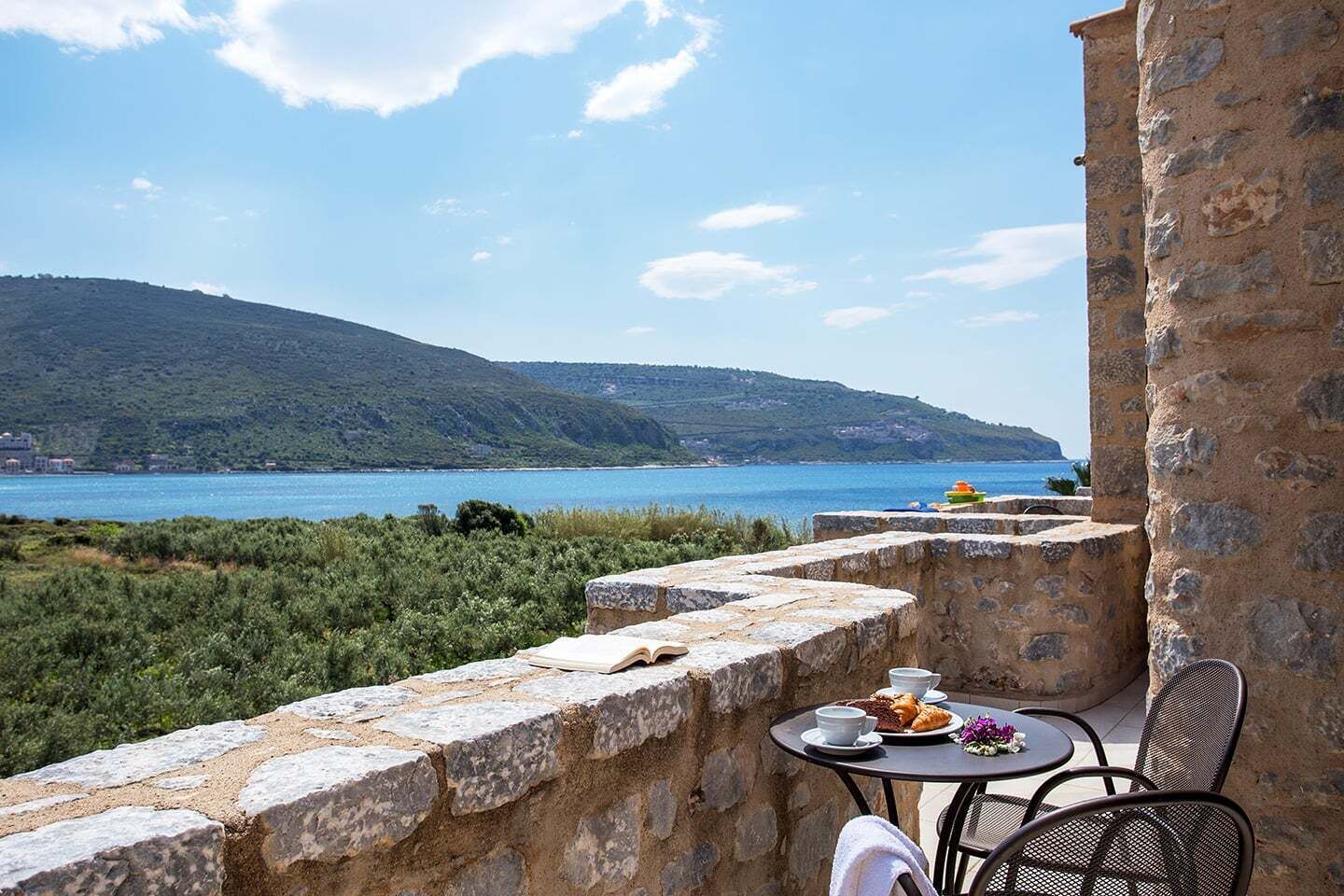 A stone balcony with a table and chairs, overlooking a body of water and a hilly landscape.