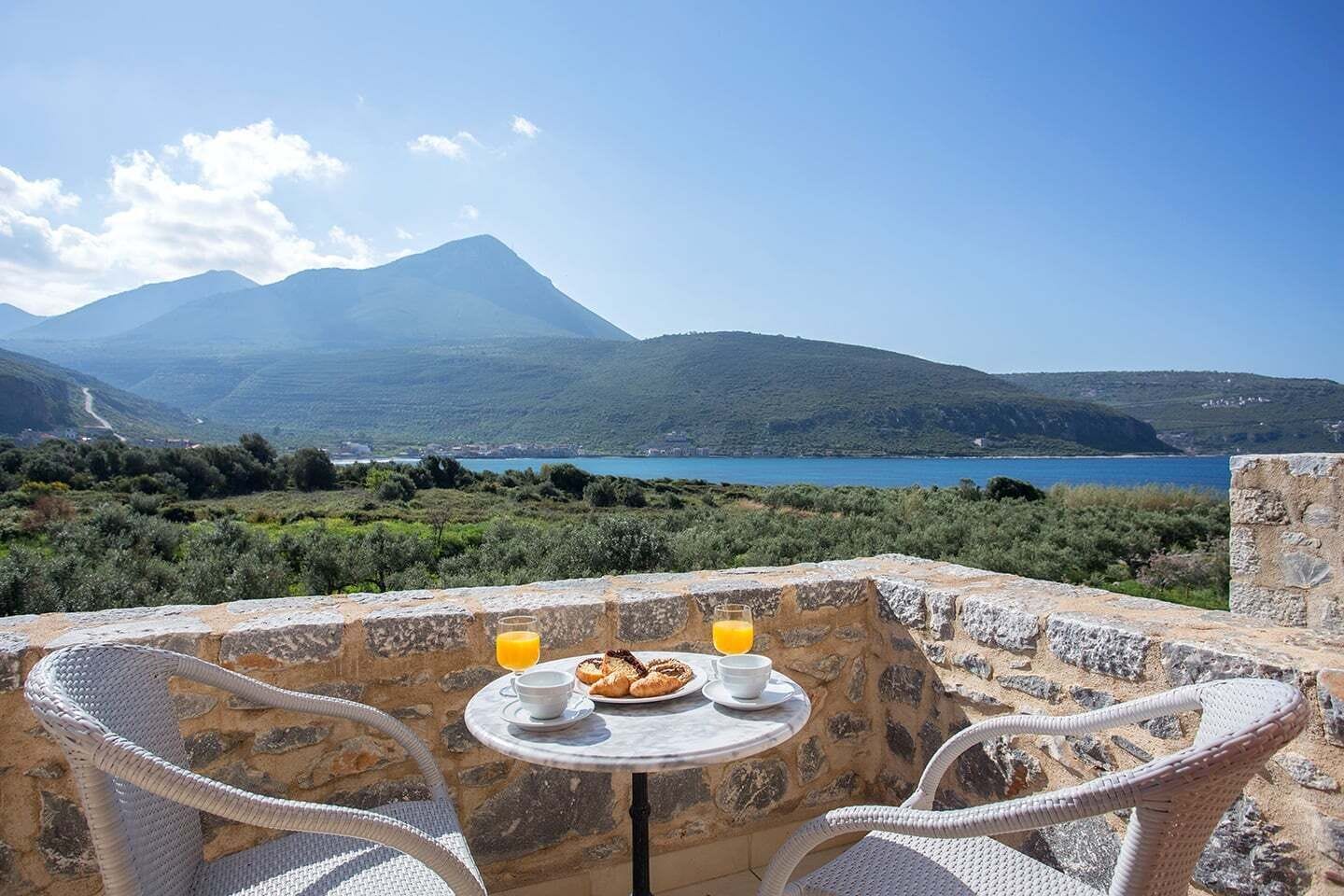 Une table en pierre, dressée avec des aliments et des boissons pour le petit-déjeuner, offre une vue panoramique sur un paysage magnifique, avec des montagnes et une étendue d’eau.