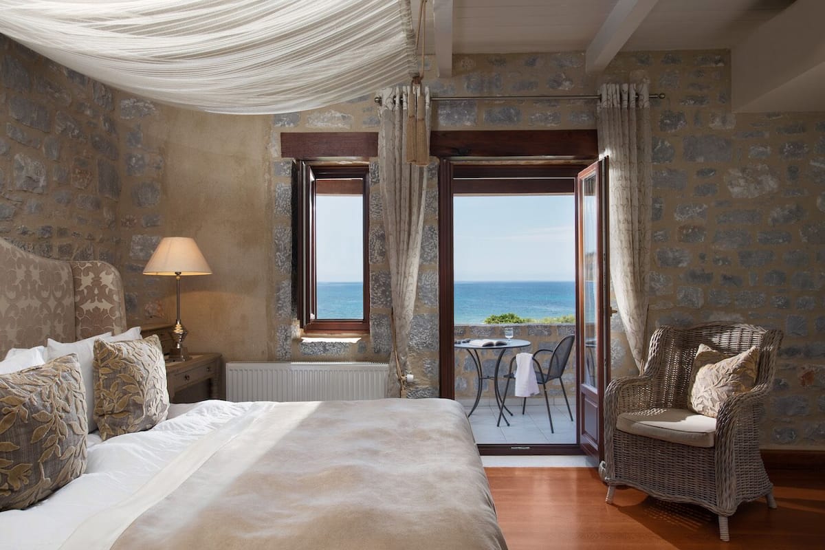Een slaapkamer met een groot bed, een nachtkastje, een lamp, een stoel en uitzicht op zee door een open deur.