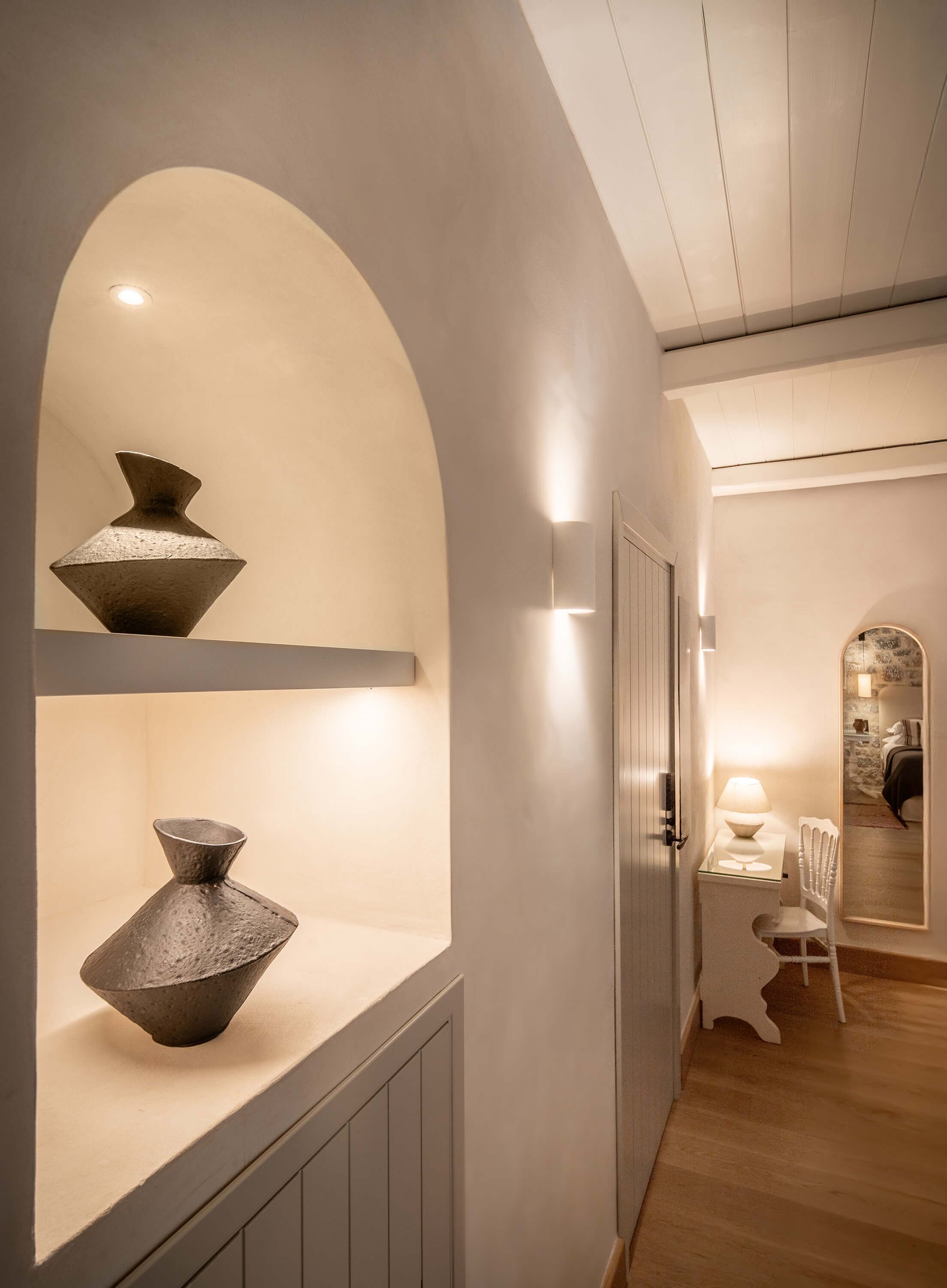 A hallway with a white wall, a wooden floor, and a small table with a lamp.