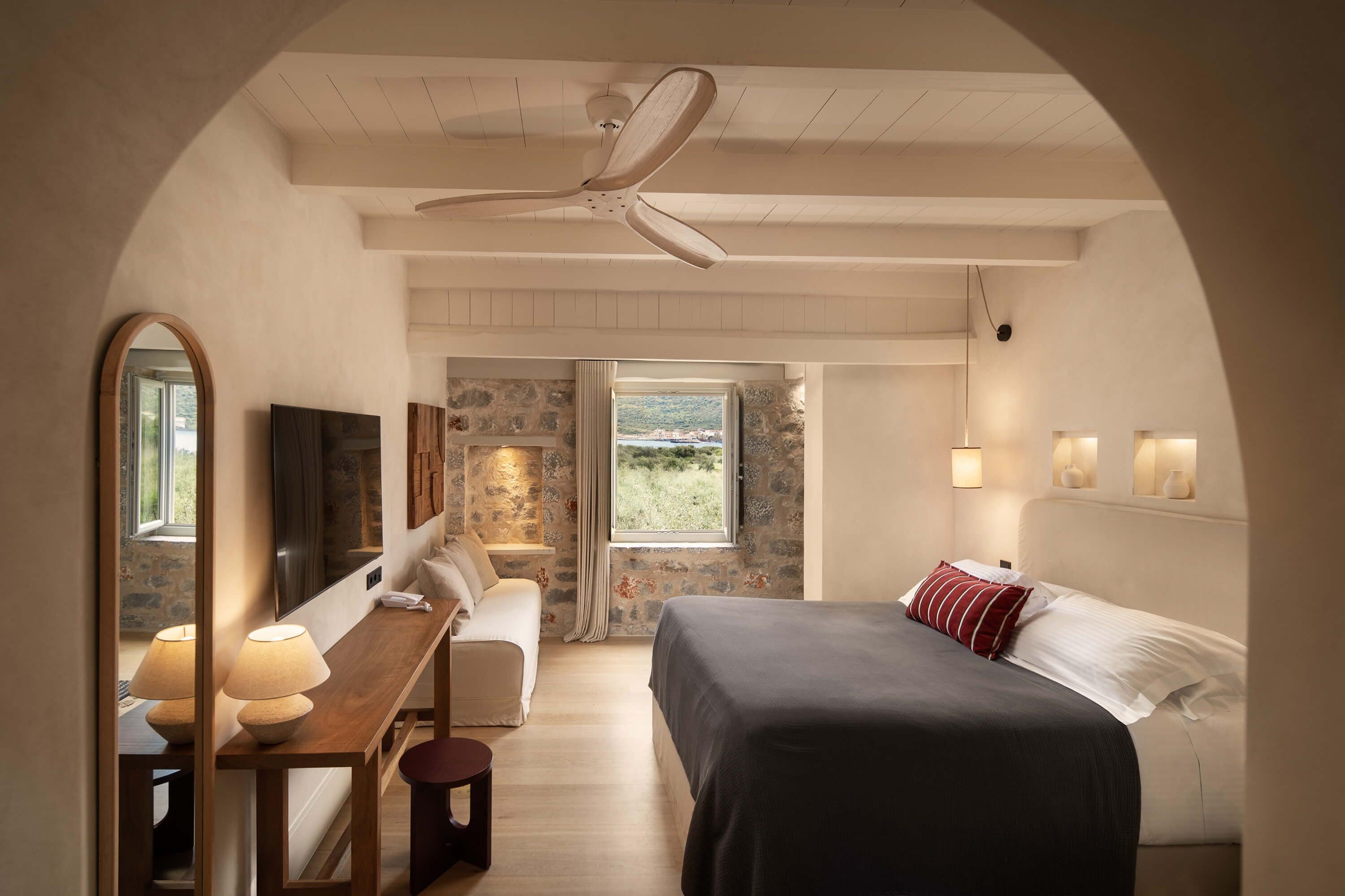 A bedroom with a bed, a desk with a lamp, a mirror, a ceiling fan, and a stone wall.