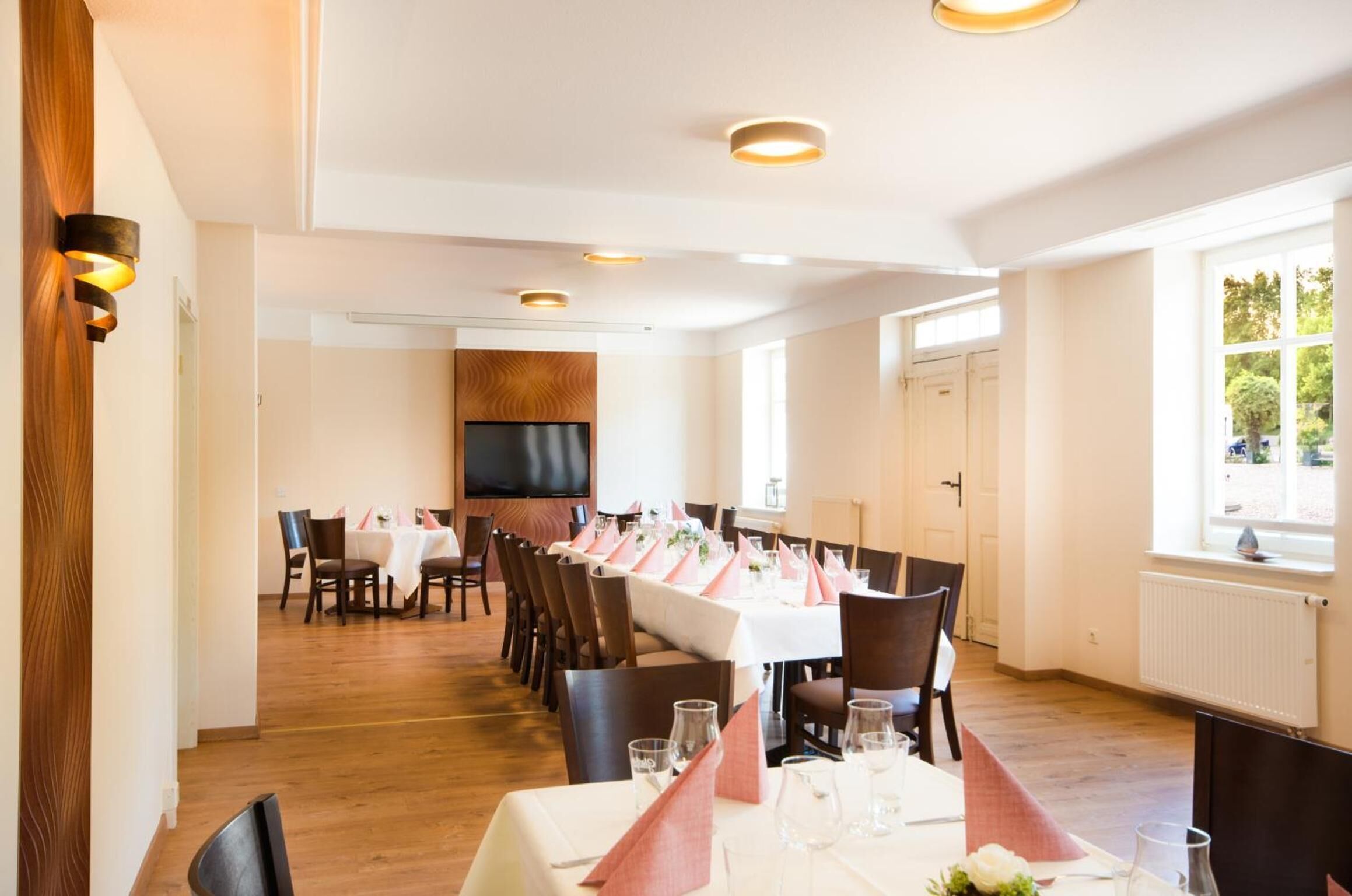 A dining area with a long table set for a meal, wooden flooring, and a TV mounted on the wall.