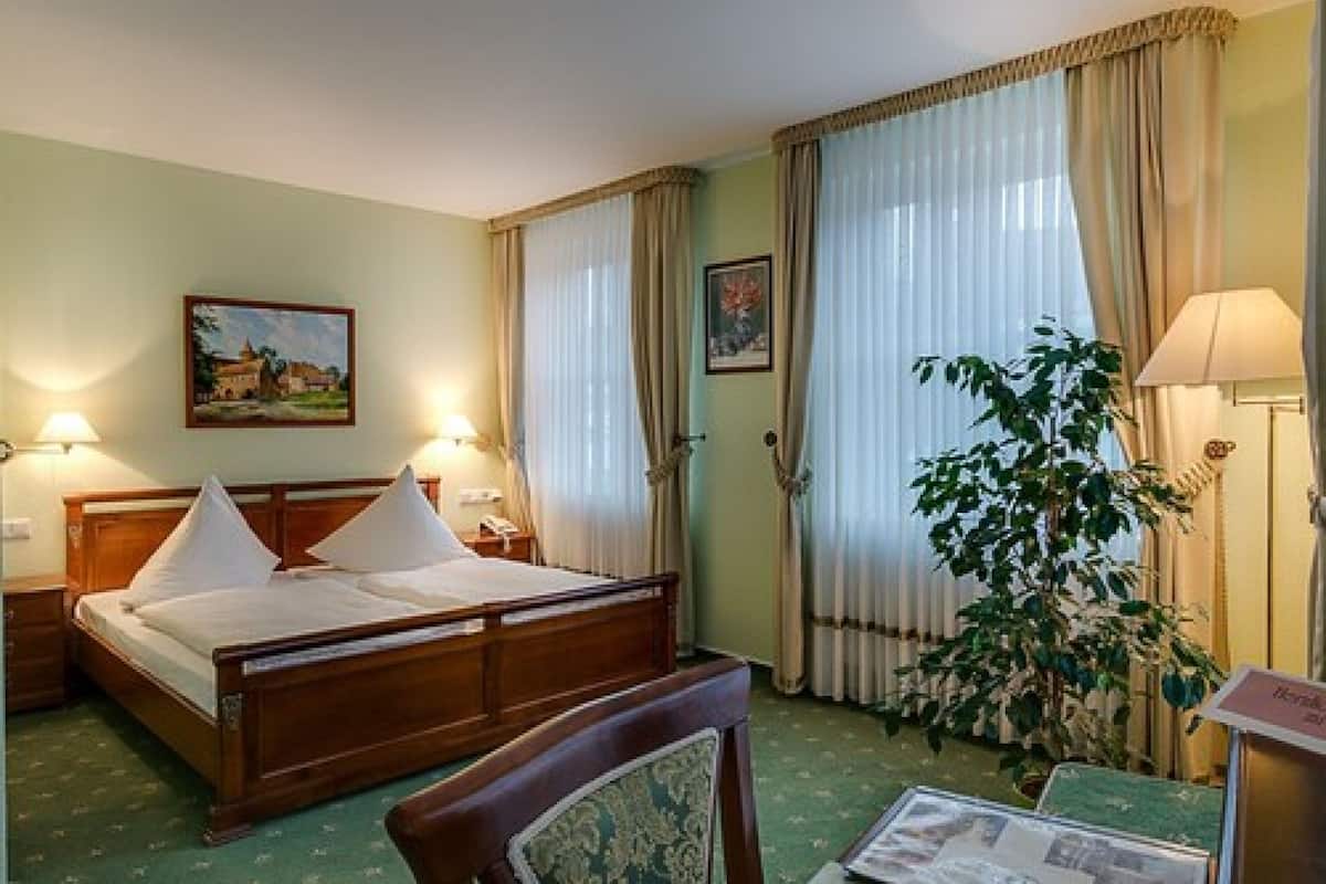 A hotel room with a bed, two bedside lamps, a painting on the wall, a window with curtains, and a plant.