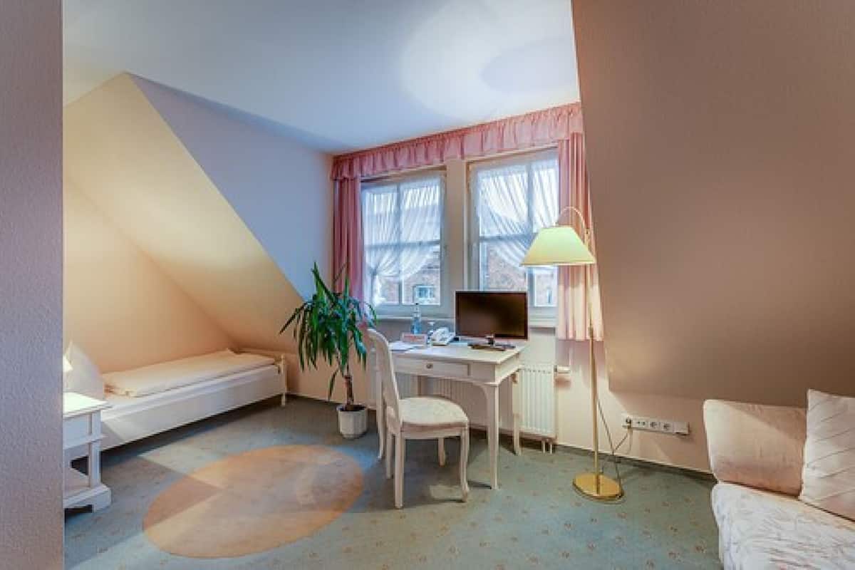 A small, cozy room with a bed, a desk, a chair, a lamp, and a window.