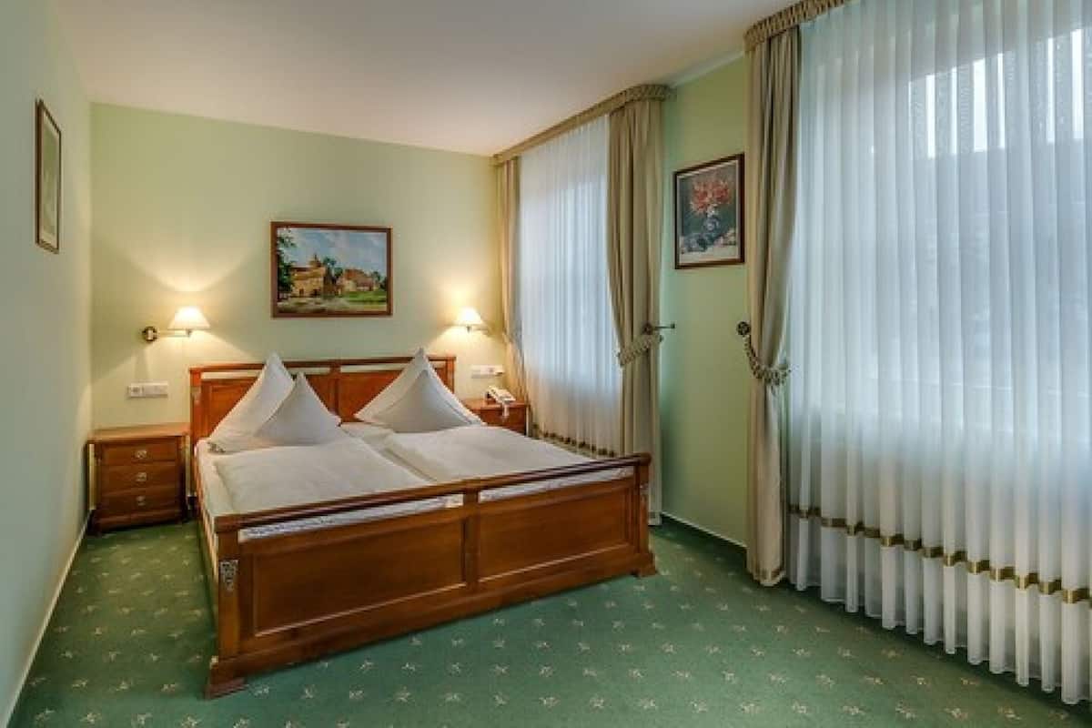 A hotel room with a wooden bed, two bedside tables, a nightstand, a painting on the wall, and a window with curtains.