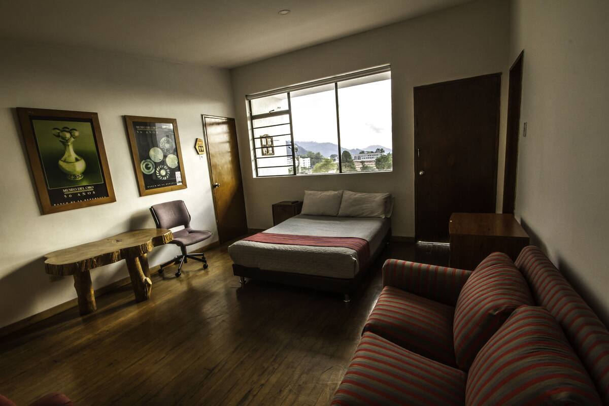 Una camera da letto con un letto, una scrivania in legno, una sedia, un divano e una finestra vista edifici e montagne.