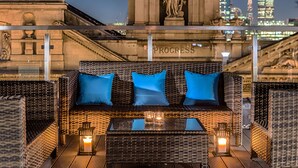 Un sofá de mimbre con cojines azules, una mesita de vidrio y velas, con un paisaje urbano de fondo.