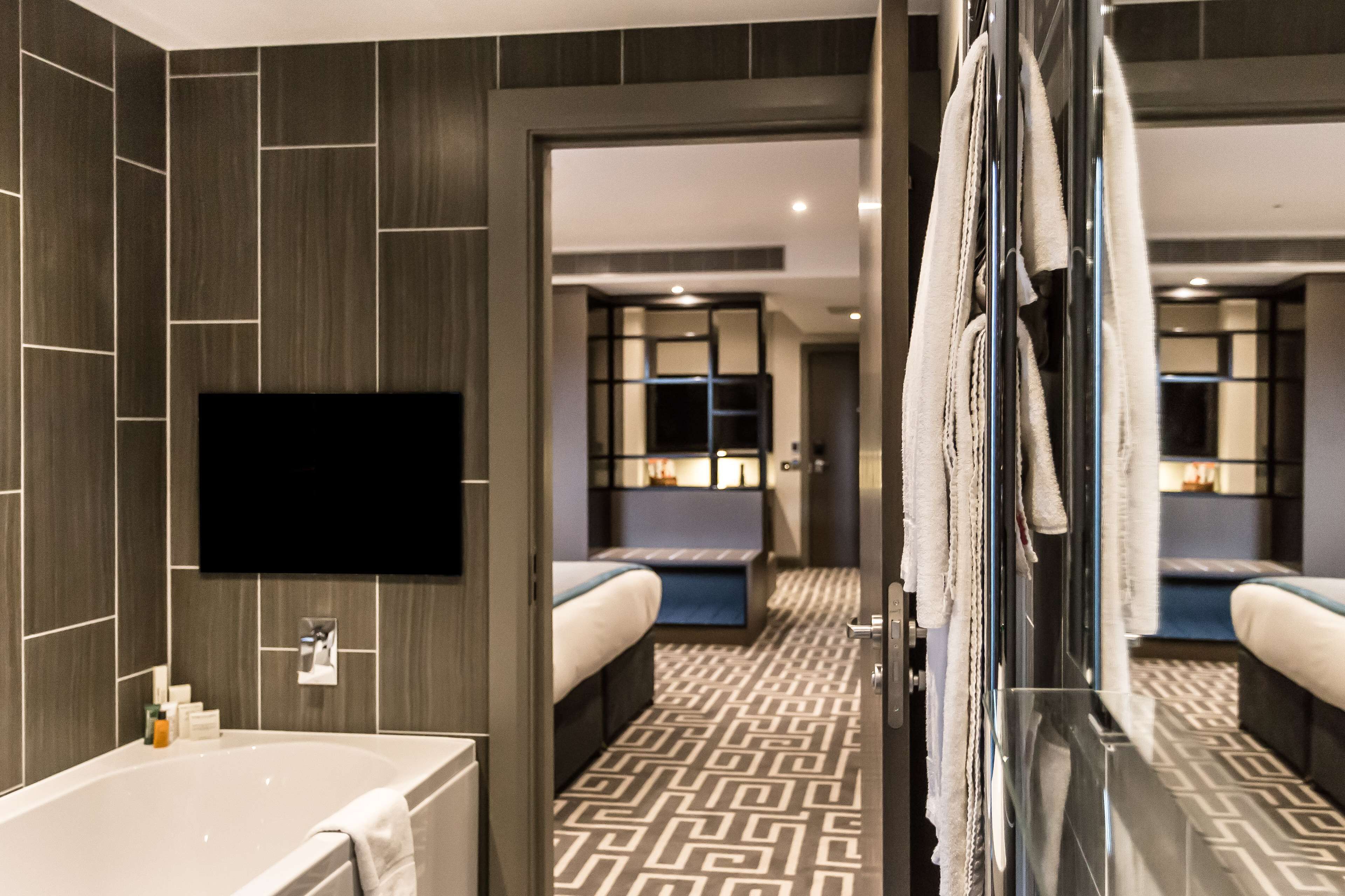 Une salle de bain d’hôtel moderne, dotée d’une grande baignoire, d’un téléviseur à écran plat fixé au mur et d’un carrelage décoratif.