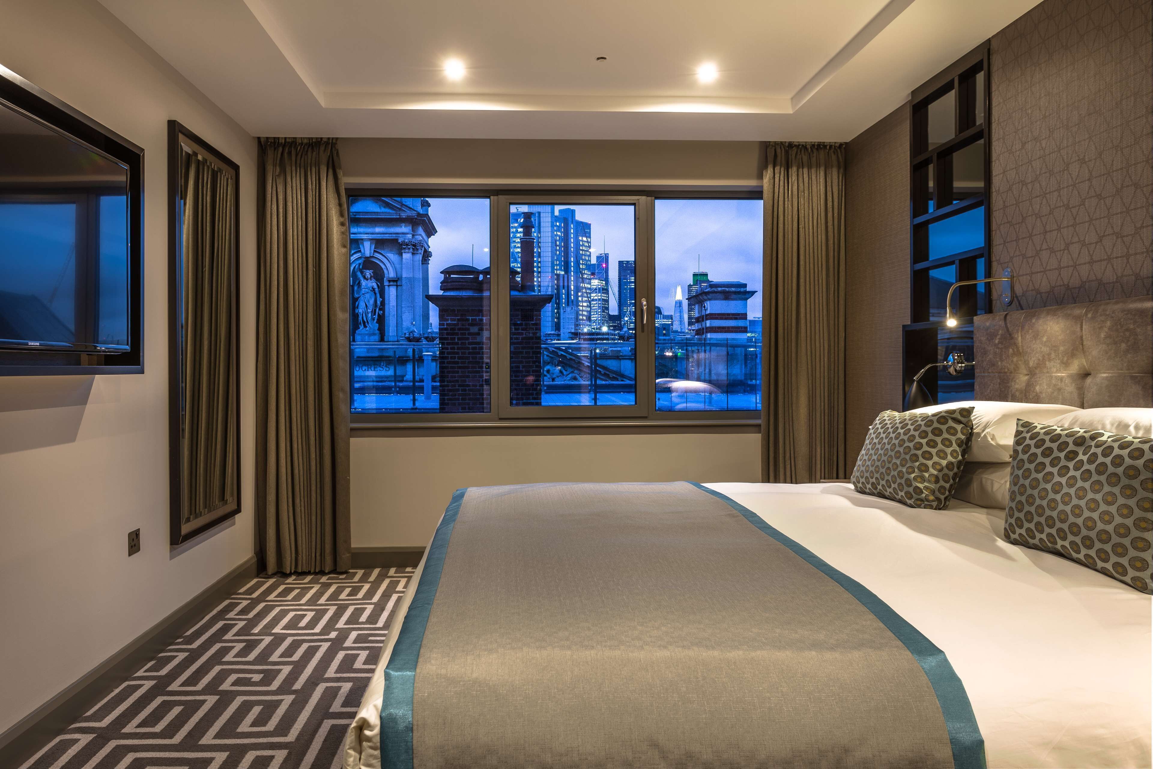 A hotel room with a large bed, a city view, a TV, and a patterned floor.