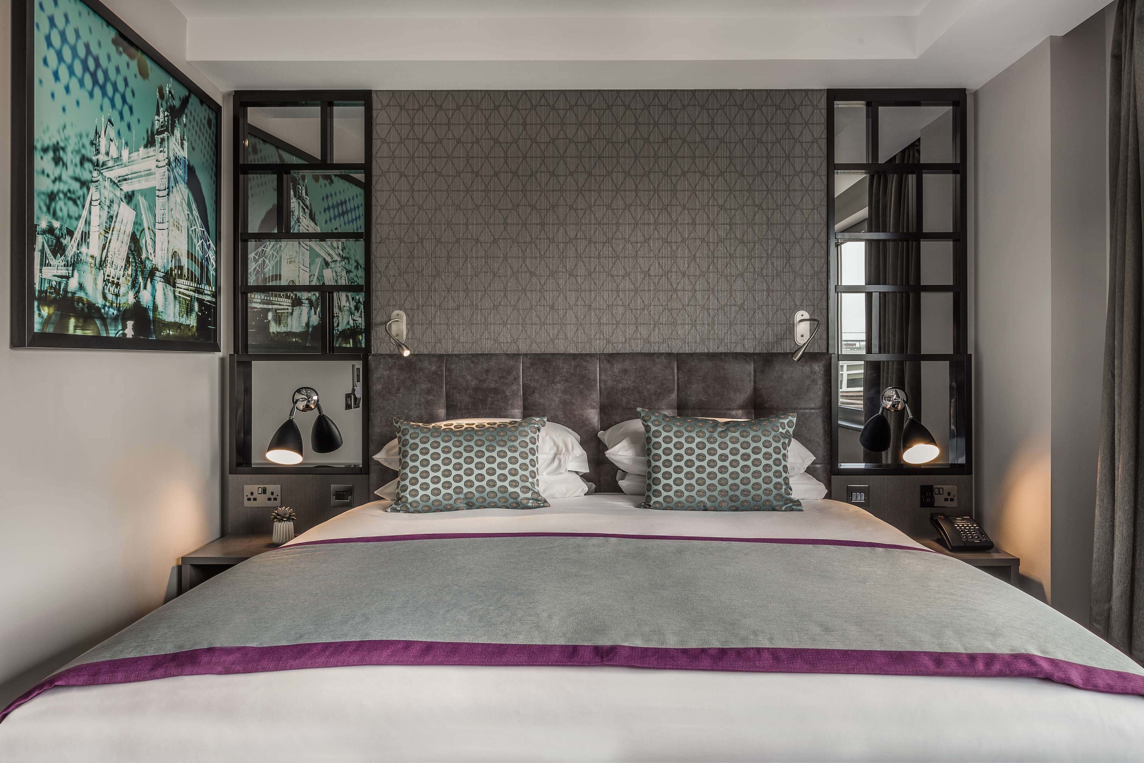 A modern hotel room with a large bed, a headboard with a geometric pattern, a nightstand with a lamp, and a wall-mounted mirror.
