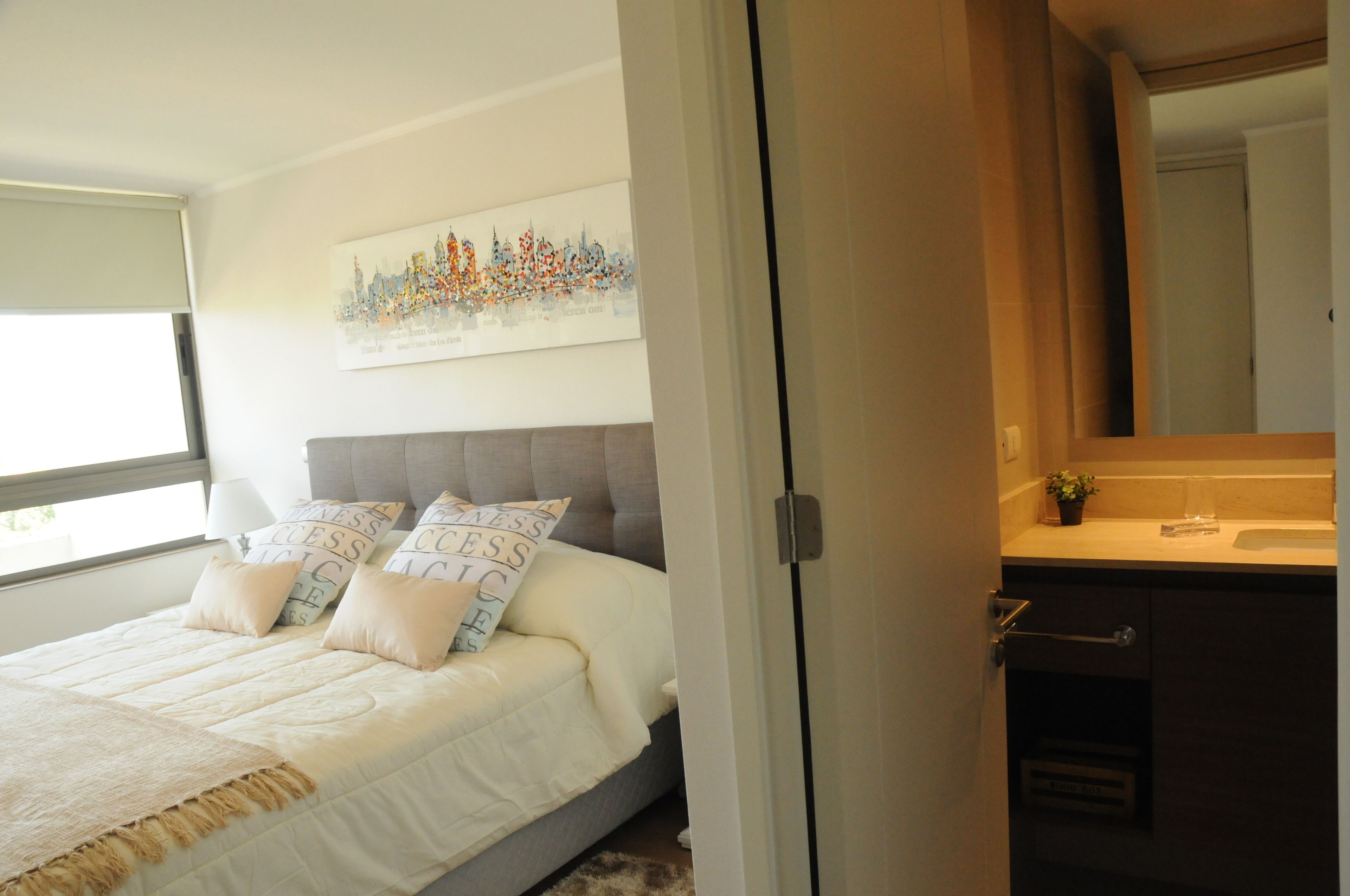 Un dormitorio con cama, almohadas, mesita de noche, ventana y baño con lavabo y espejo.