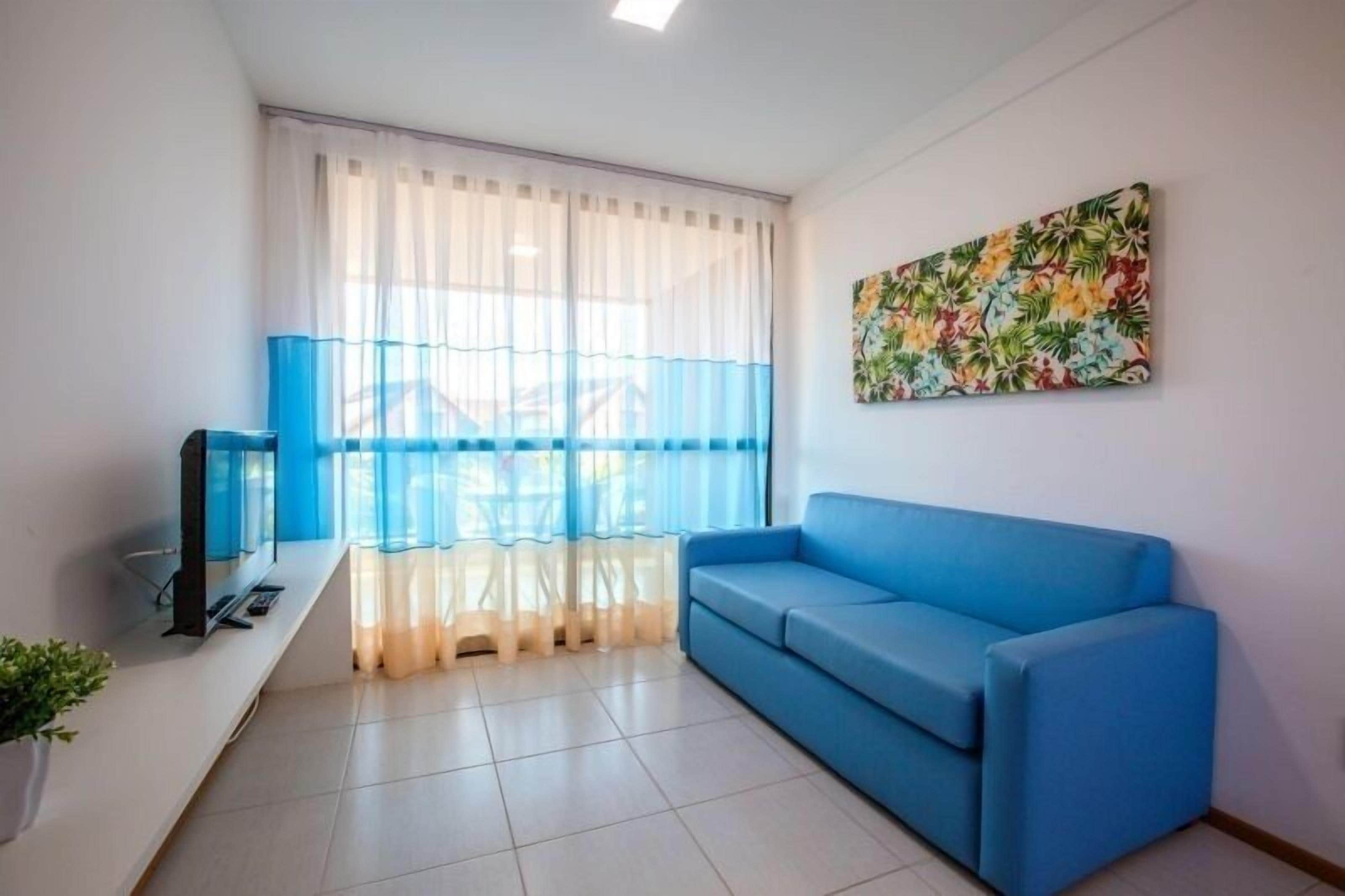 Uma sala de estar com um sofá azul, uma televisão em um móvel, uma janela com cortinas leves e um quadro com tema tropical na parede.