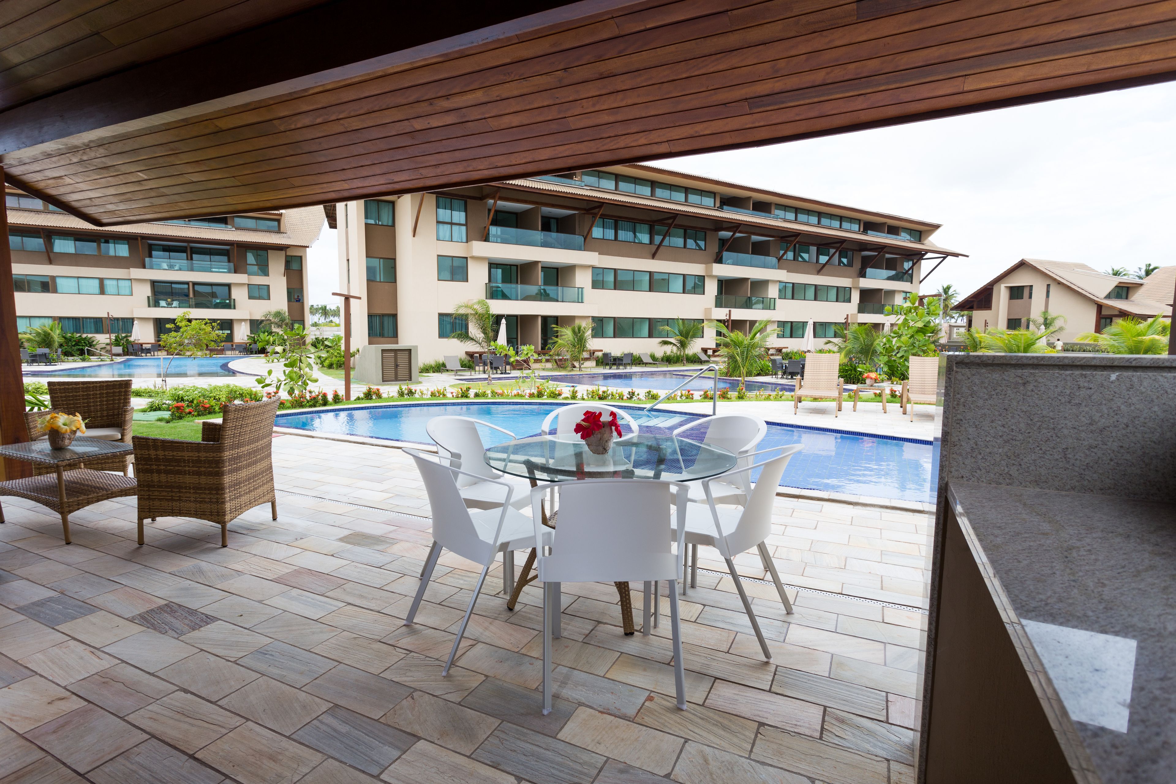 A poolside area with a glass table and chairs, a wicker chair, and a view of a multi-story building with balconies.