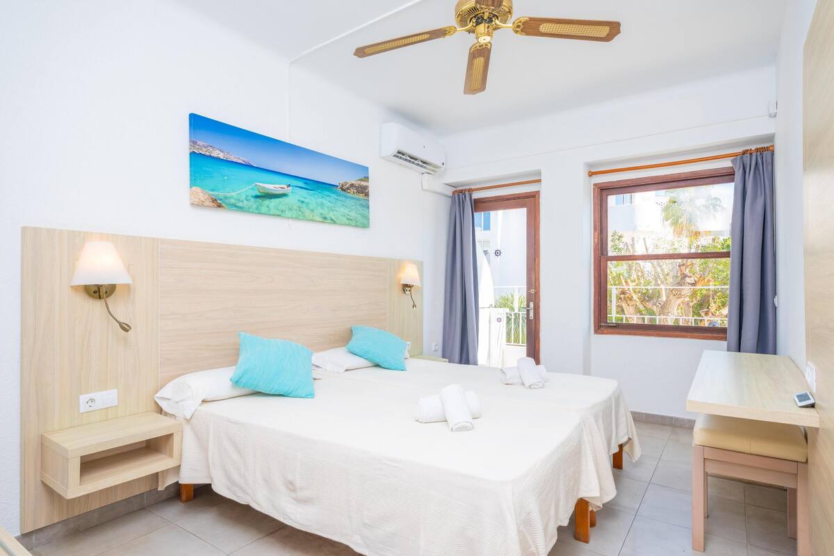 A hotel room with a bed, two bedside tables, a desk, a ceiling fan, an air conditioning unit, a window with curtains, and a painting of a beach scene.