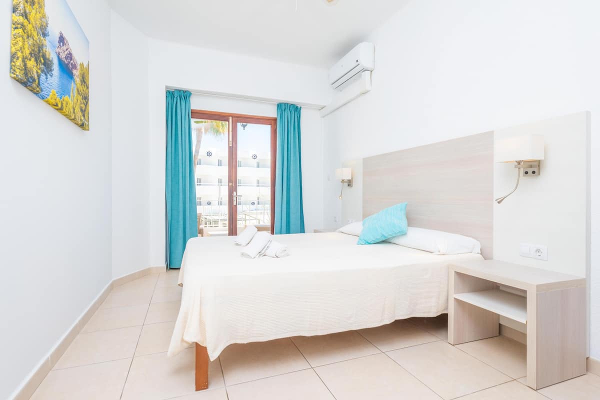 A hotel room with a bed, a bedside table, a wall-mounted lamp, and a window with curtains.