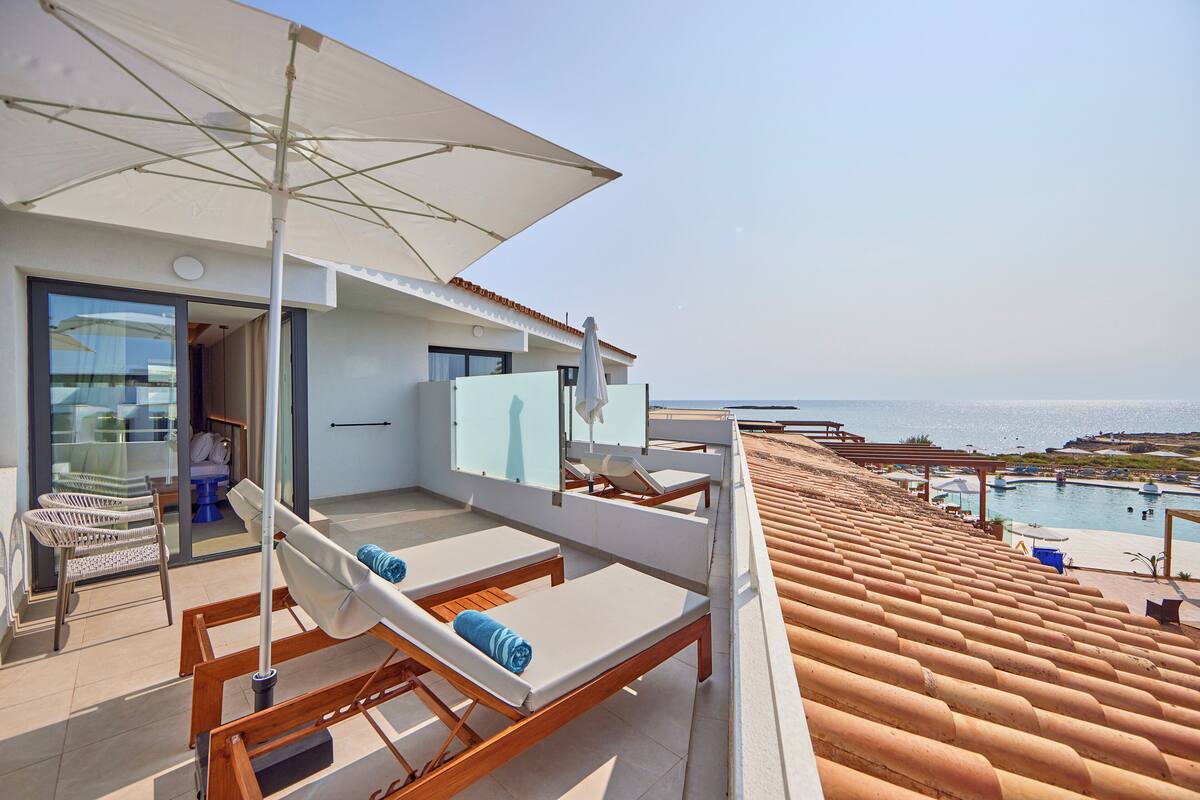 A rooftop terrace with lounge chairs, a large umbrella, and a view of the sea.