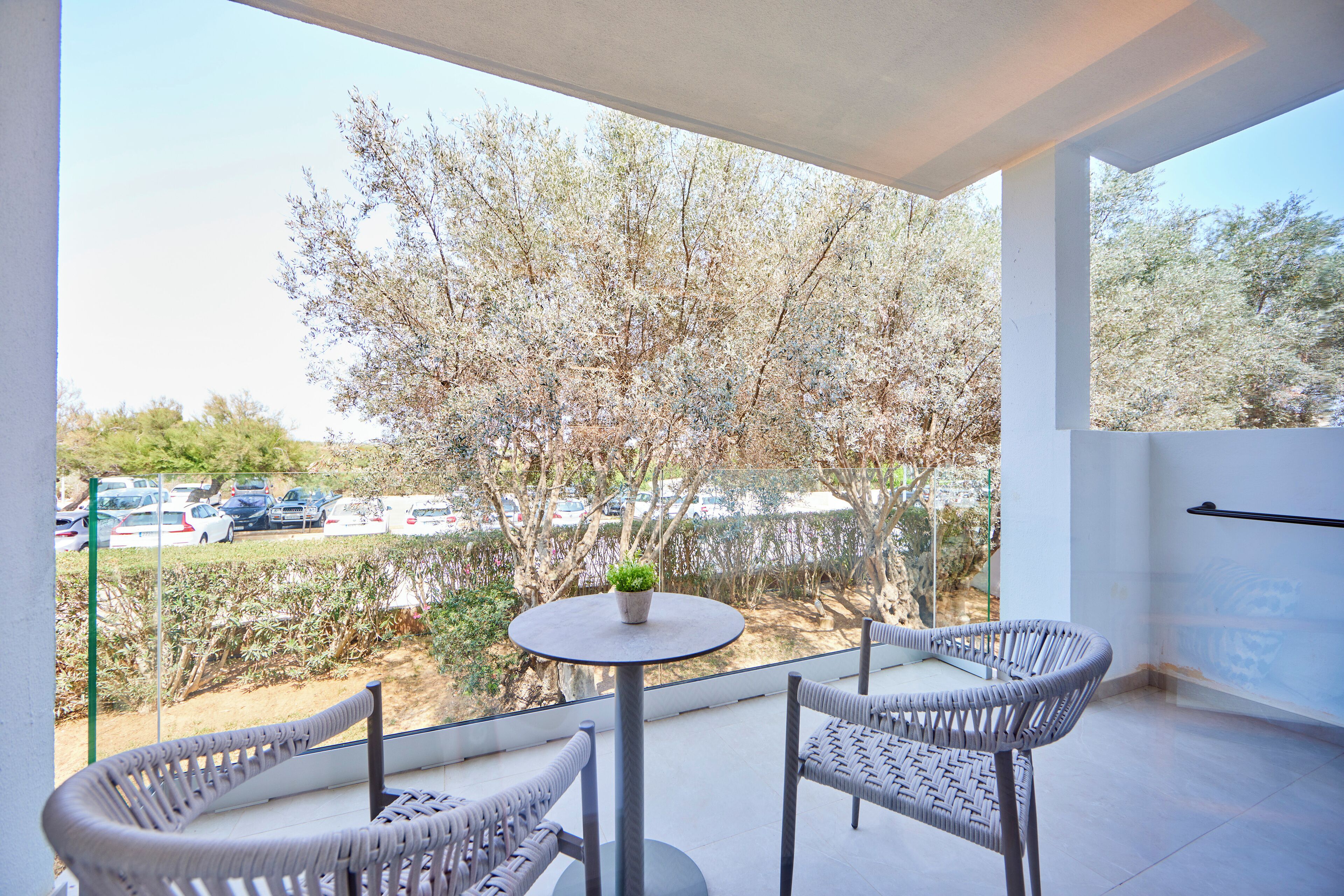 A balcony with two wicker chairs, a small round table with a potted plant, and a view of a parking lot and trees.