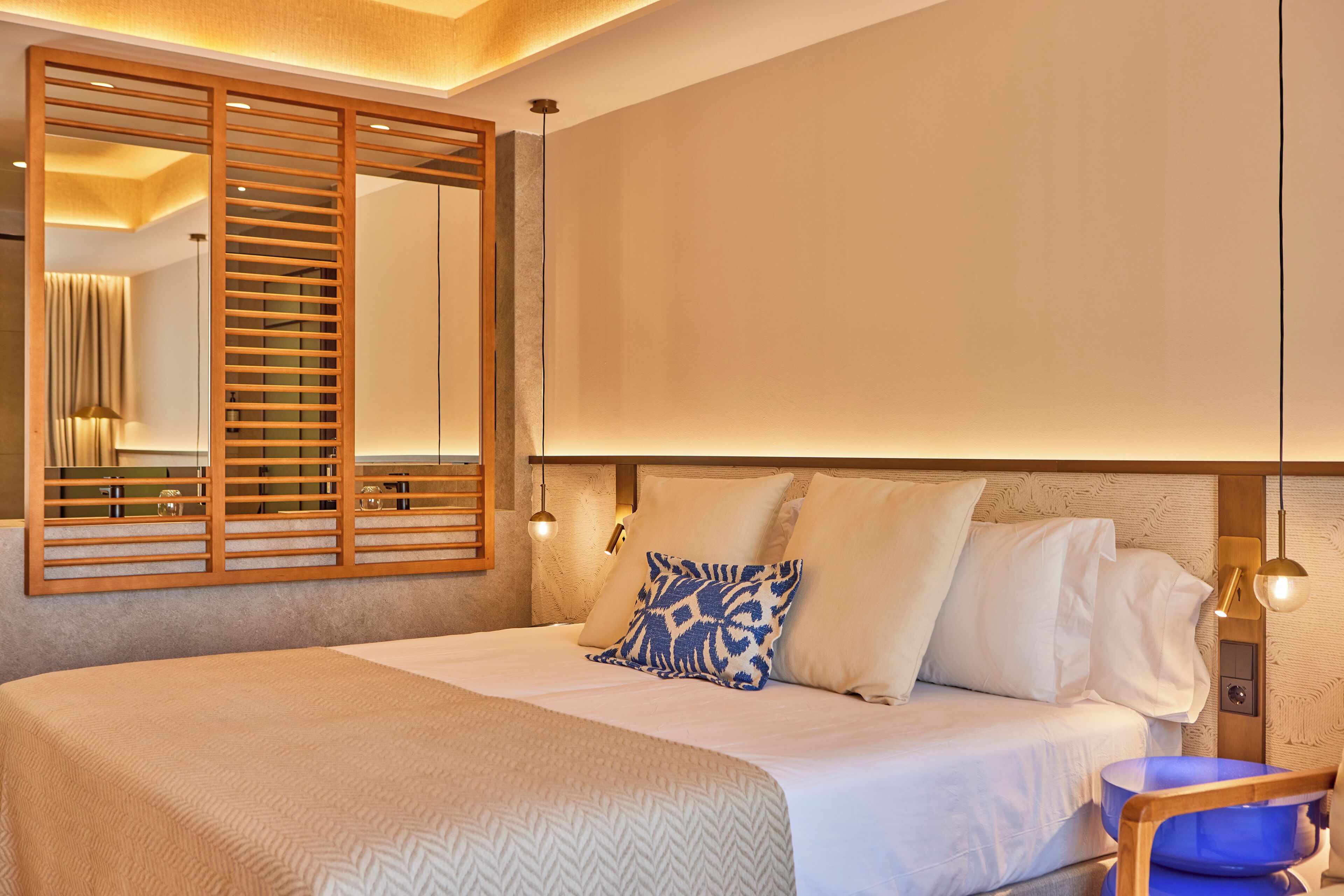 A neatly made bed with a patterned bedspread, a large mirror with wooden slats, and a bedside lamp.