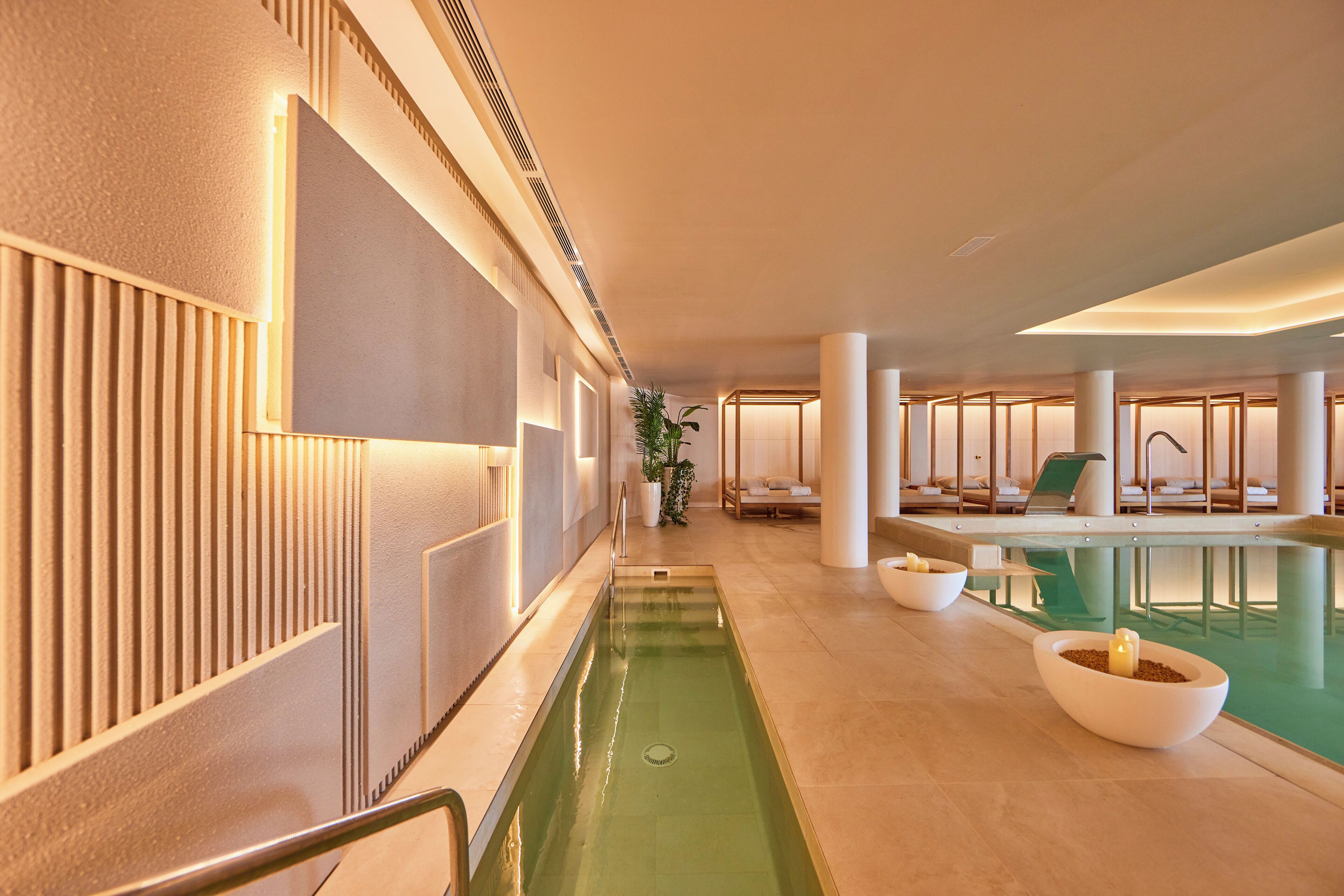 Un área de piscina interior moderna con una piscina larga y estrecha, sillas de descanso y una zona de estar con vistas a la piscina.