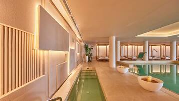 Un área de piscina interior moderna con una piscina larga y estrecha, sillas de descanso y una zona de estar con vistas a la piscina.